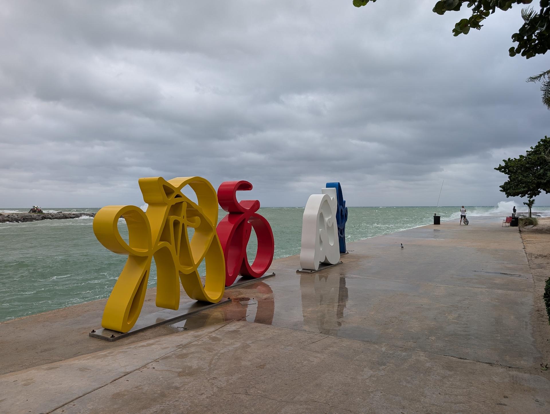 Colorful abstract sculptures on a promenade by the ocean under cloudy sky.