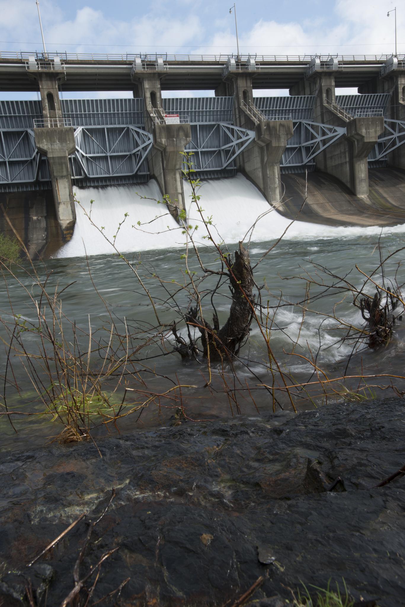 A large concrete dam releases water, with dark rocks in the foreground.