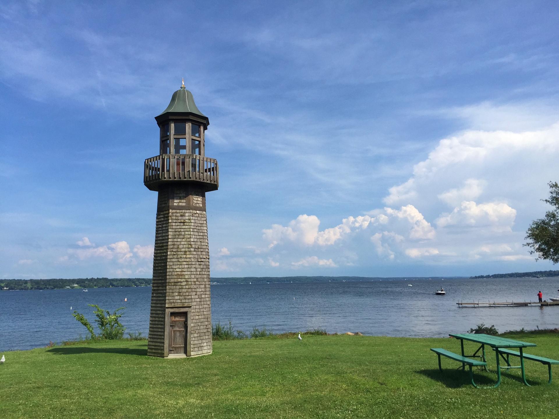 A shingled tower stands by a lake with a picnic table under a bright, cloudy sky.