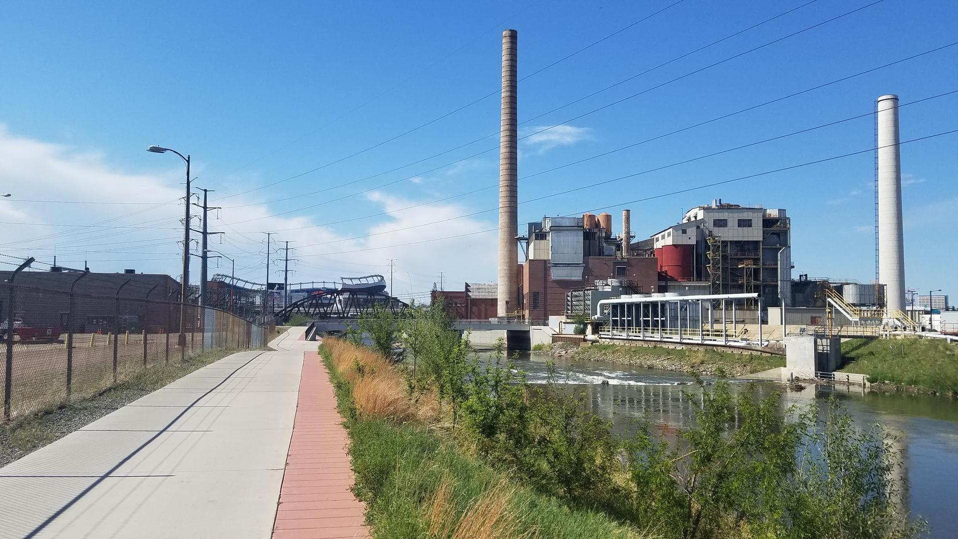Industrial buildings with tall smokestacks next to a river and path under a clear sky.