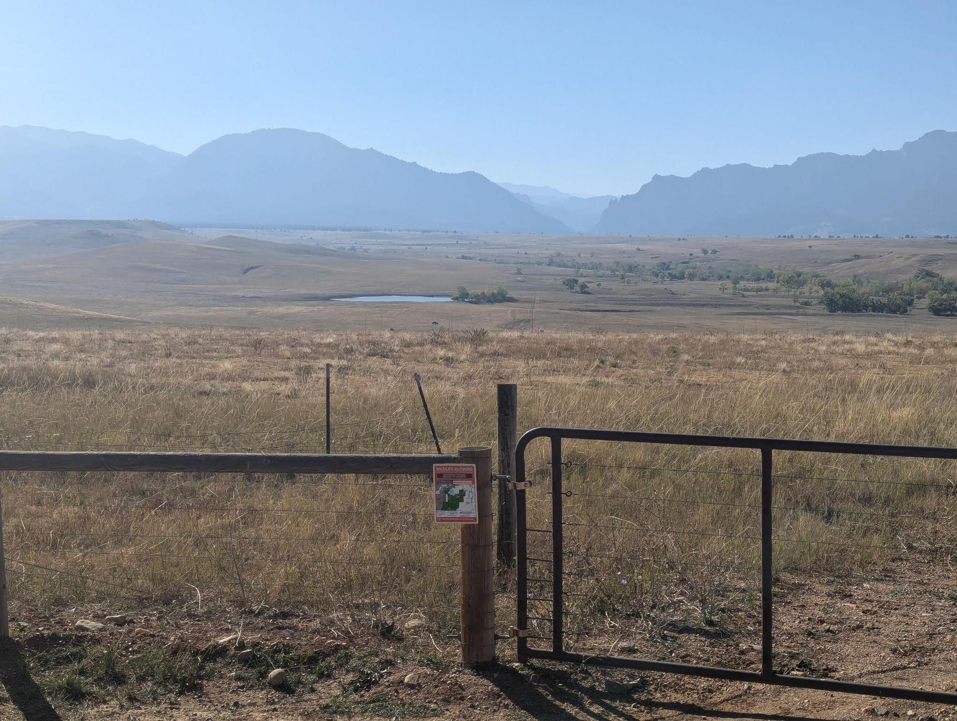 A metal gate and wooden fence in a vast dry field with hazy mountains.