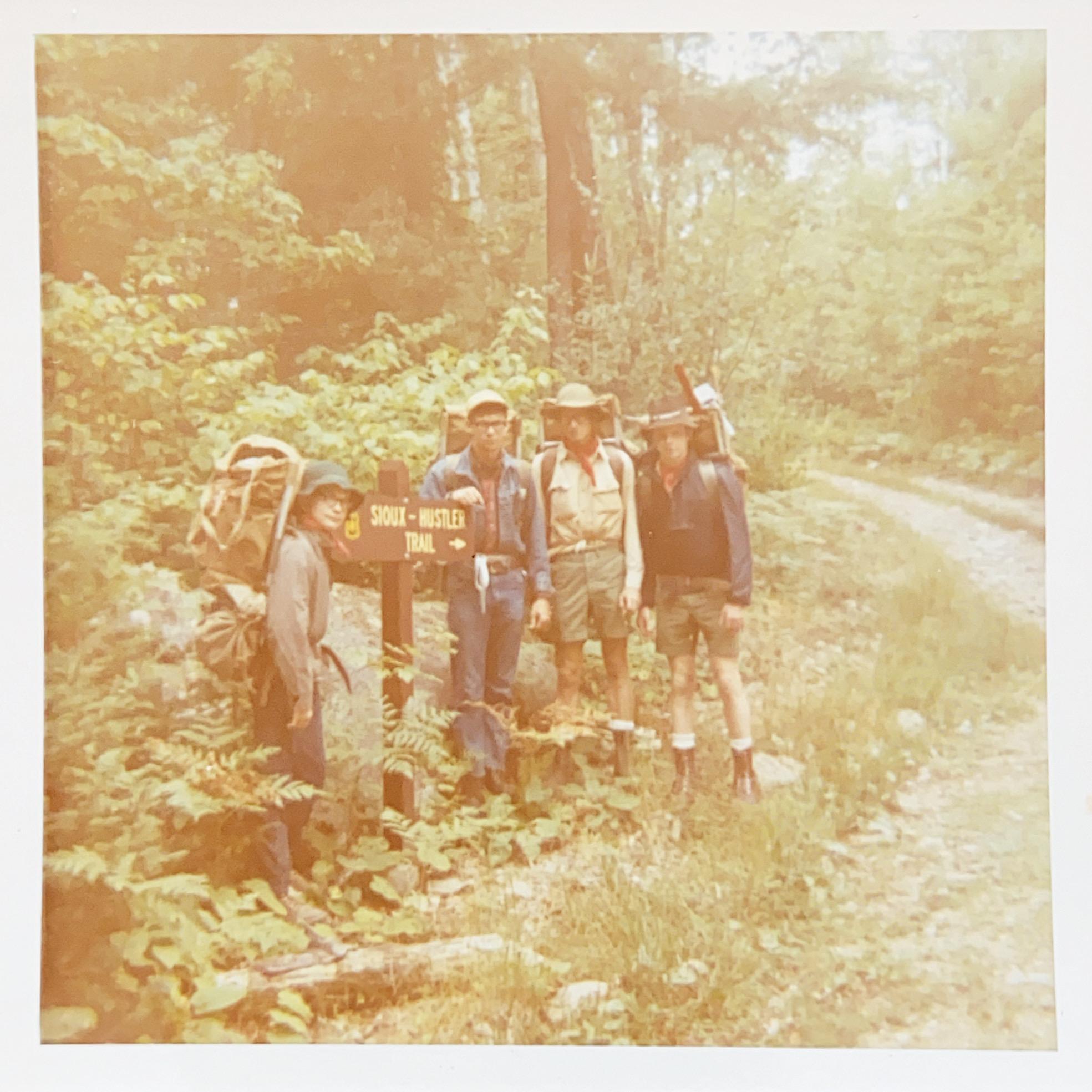 Four hikers with backpacks pose by a trail sign in bright, faded light.