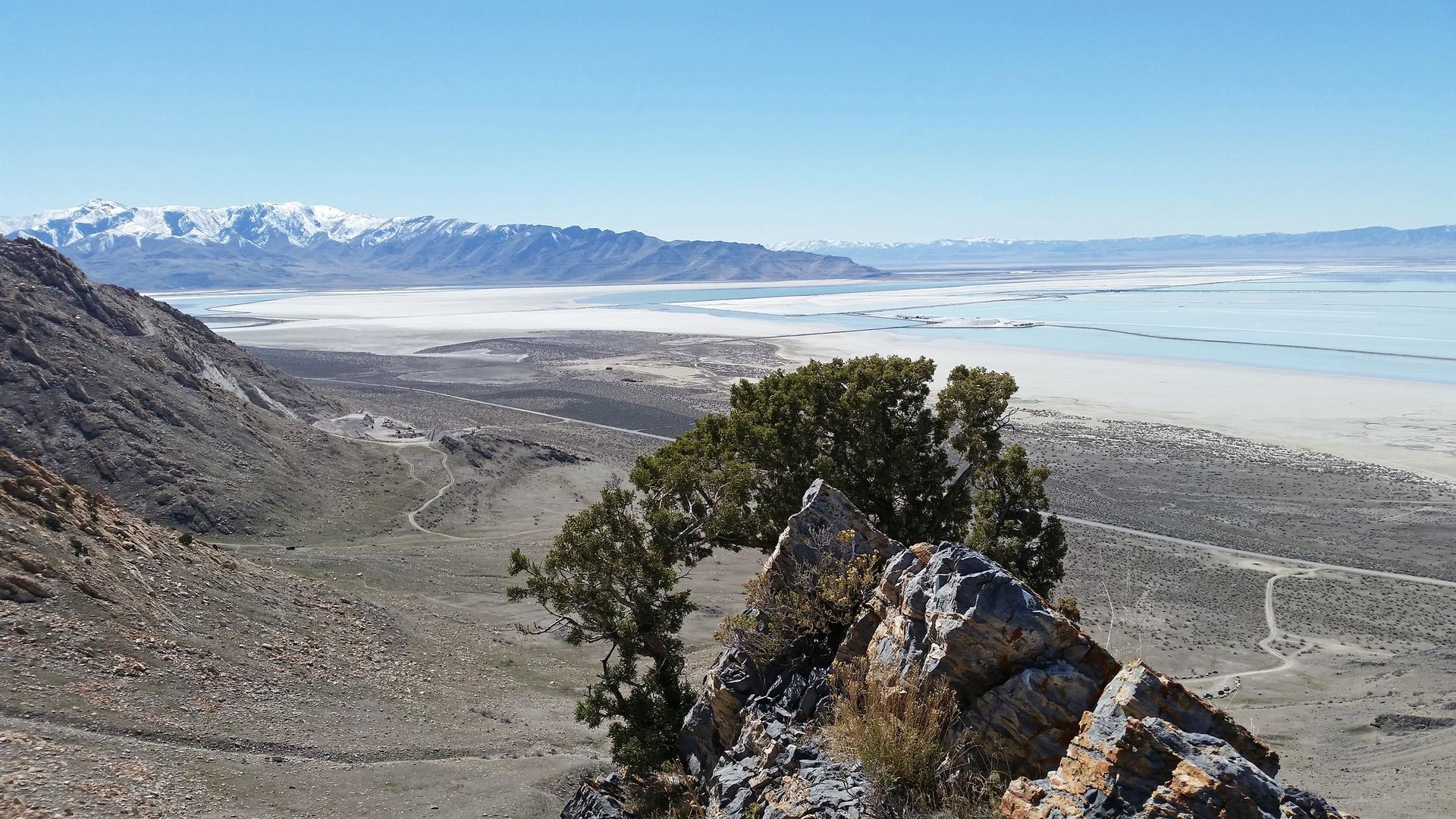 Panoramic view of a vast dry lakebed, water, and snow-capped mountains from a rocky peak.