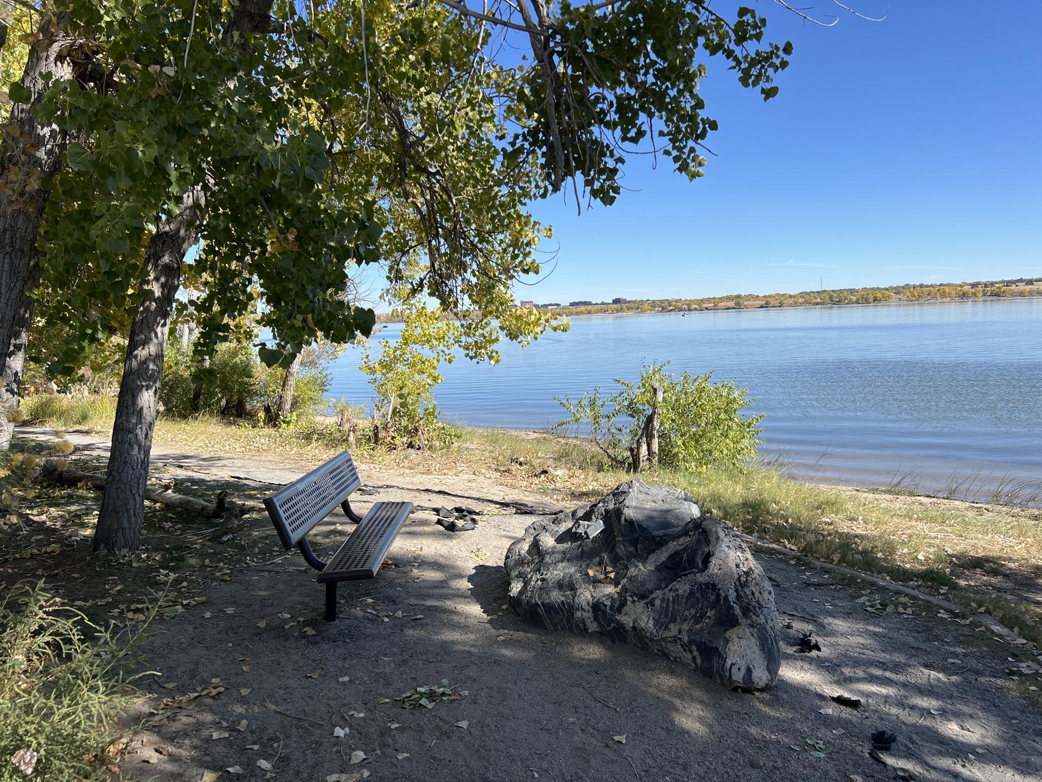 A wooden bench and rock by a calm lake under a bright blue sky.