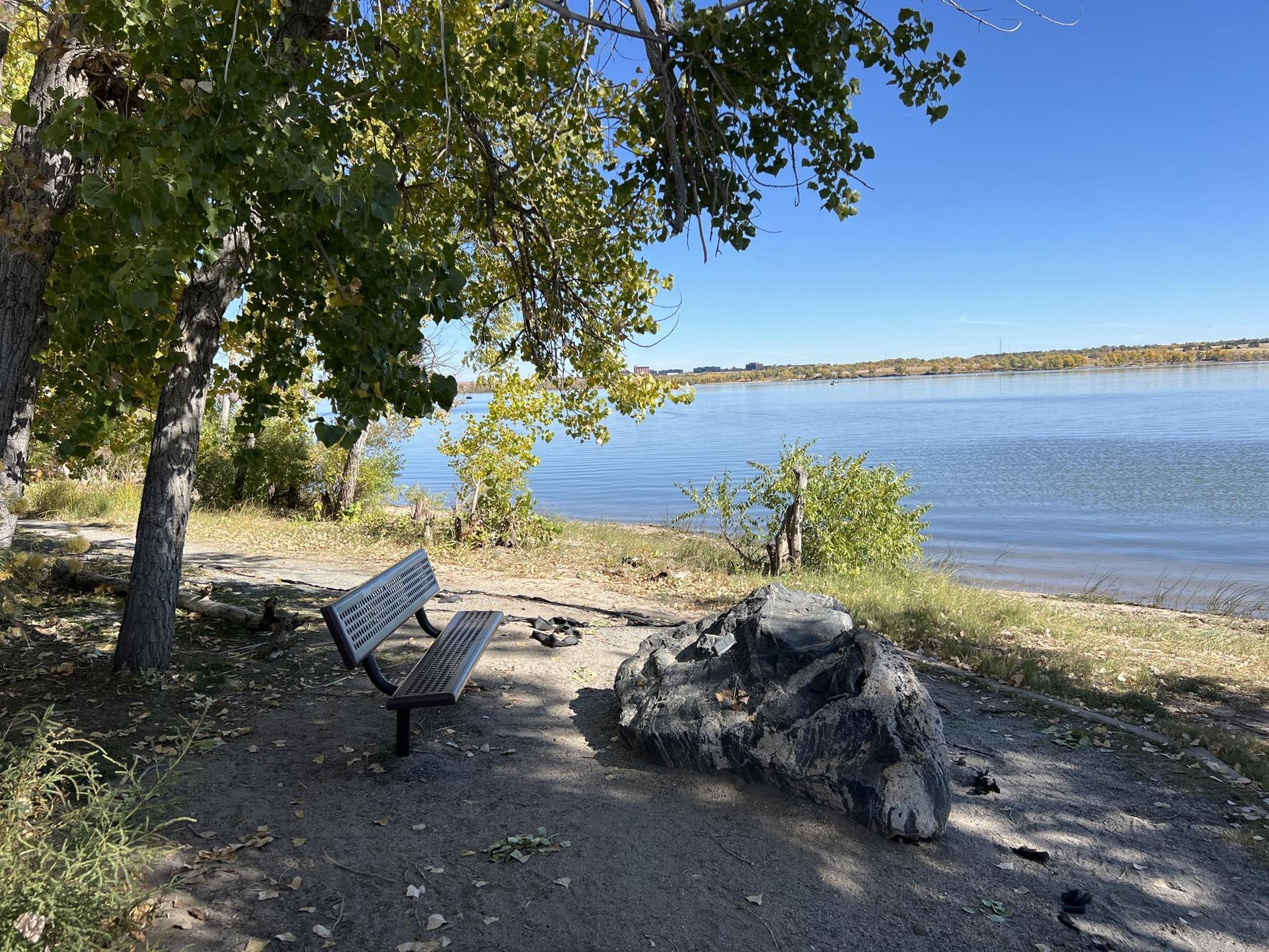 A wooden bench and rock by a calm lake under a bright blue sky.