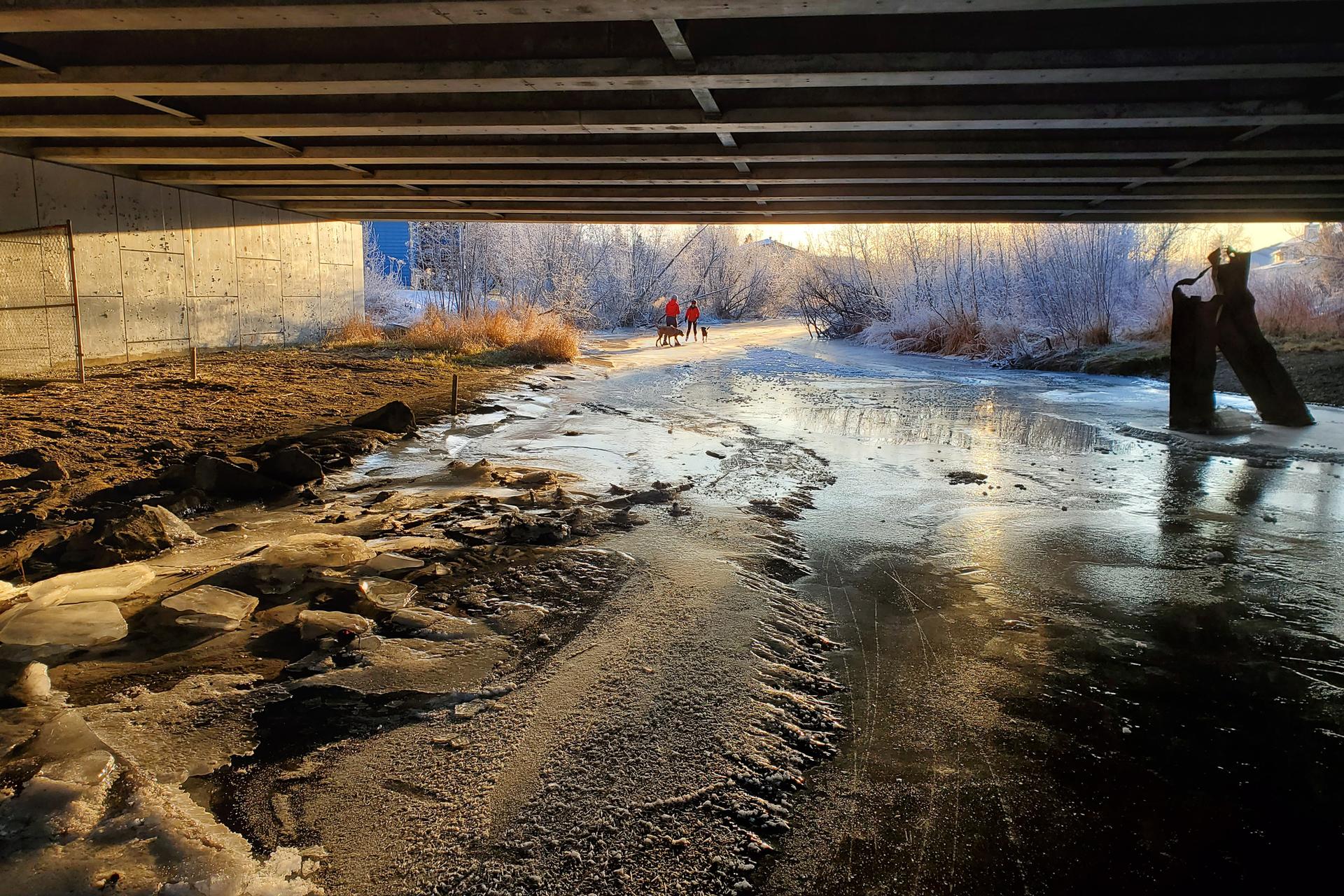 Icy river flowing under a bridge with two people walking in golden winter light.