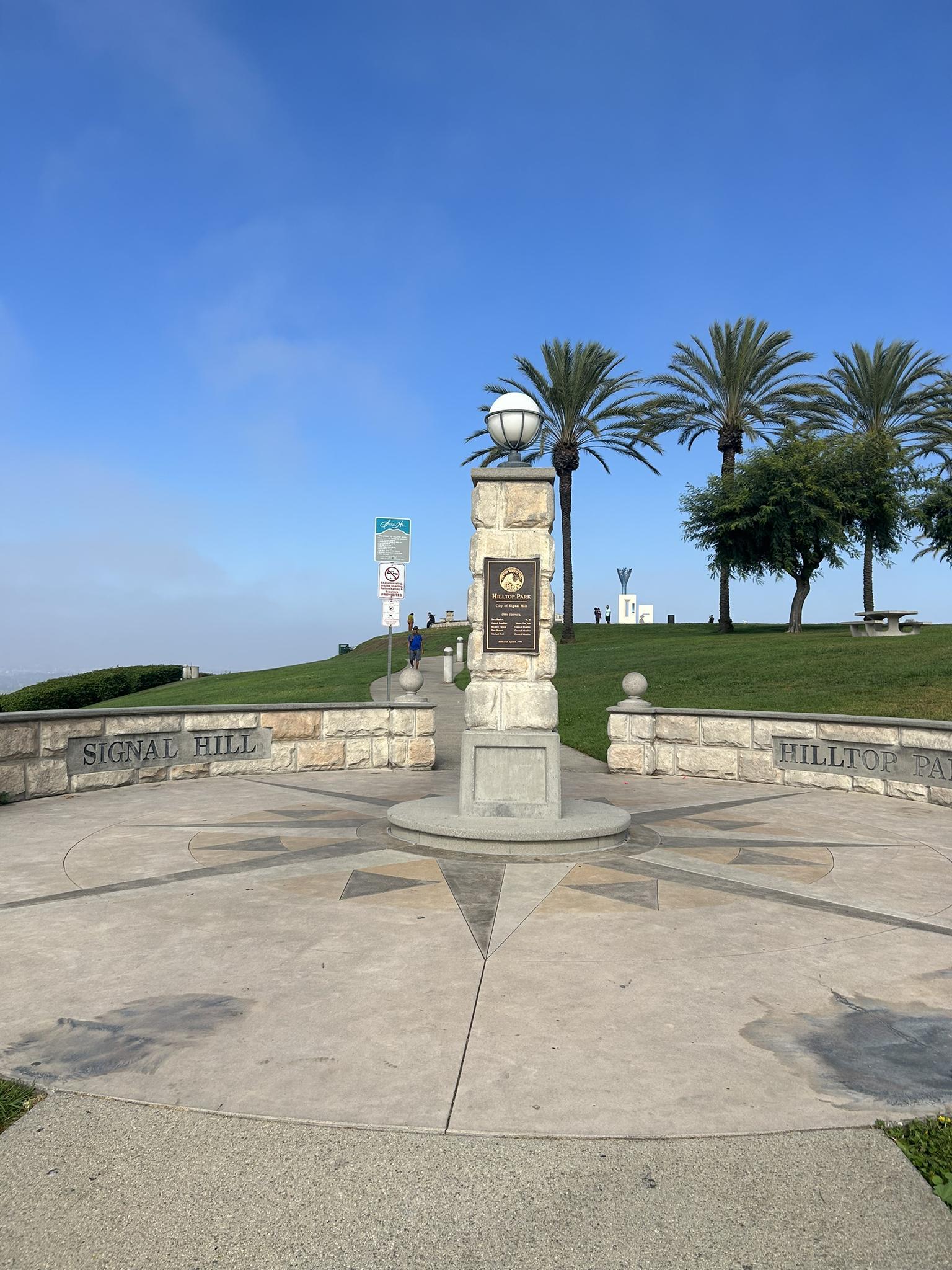 Stone monument on a paved plaza with palm trees under a clear sky.