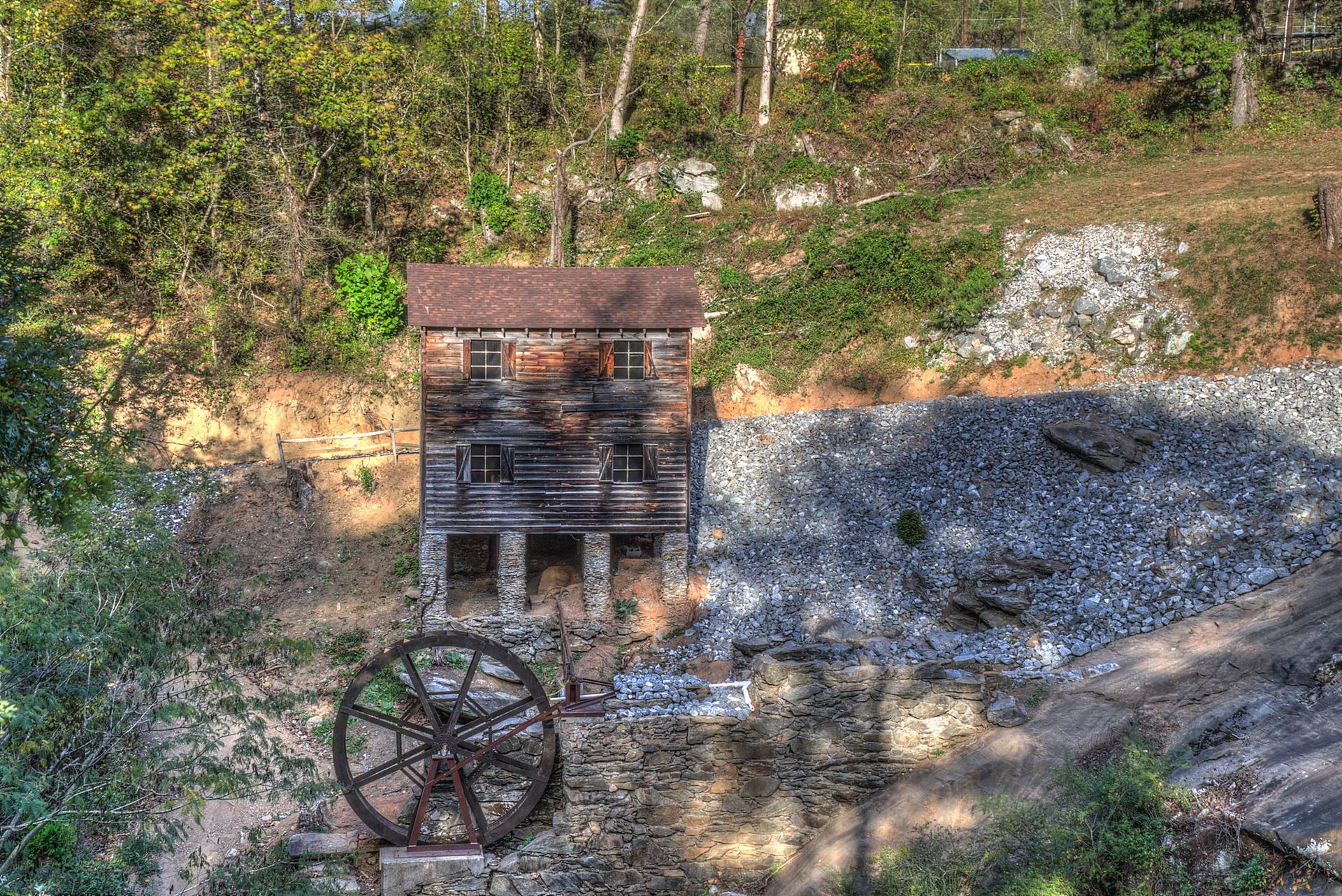 Rustic wooden building and large metal water wheel on a sunny hillside.