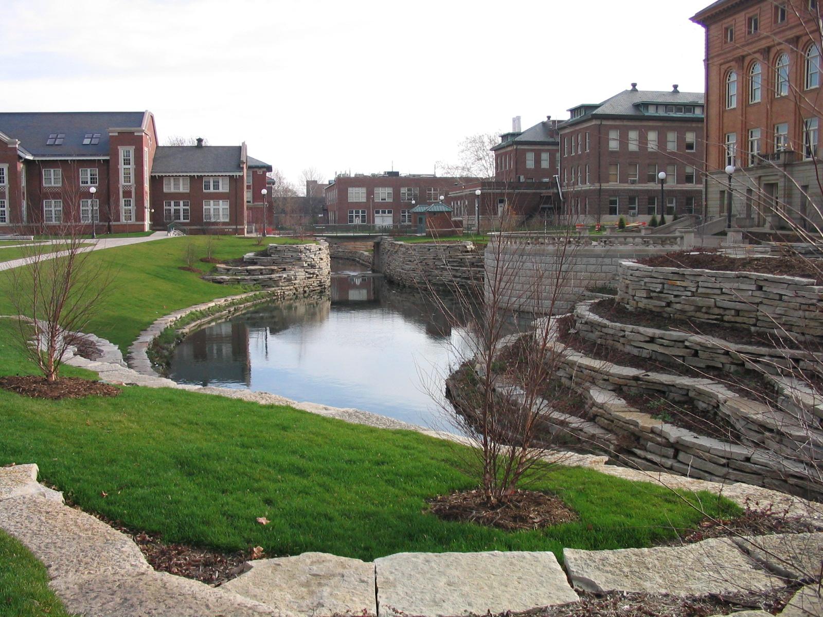 A canal flows through a landscaped urban park with brick buildings under overcast skies.