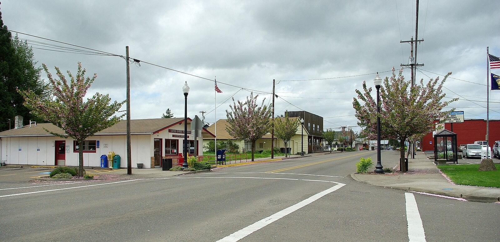 A quiet town intersection with small buildings, young trees, and an overcast sky.