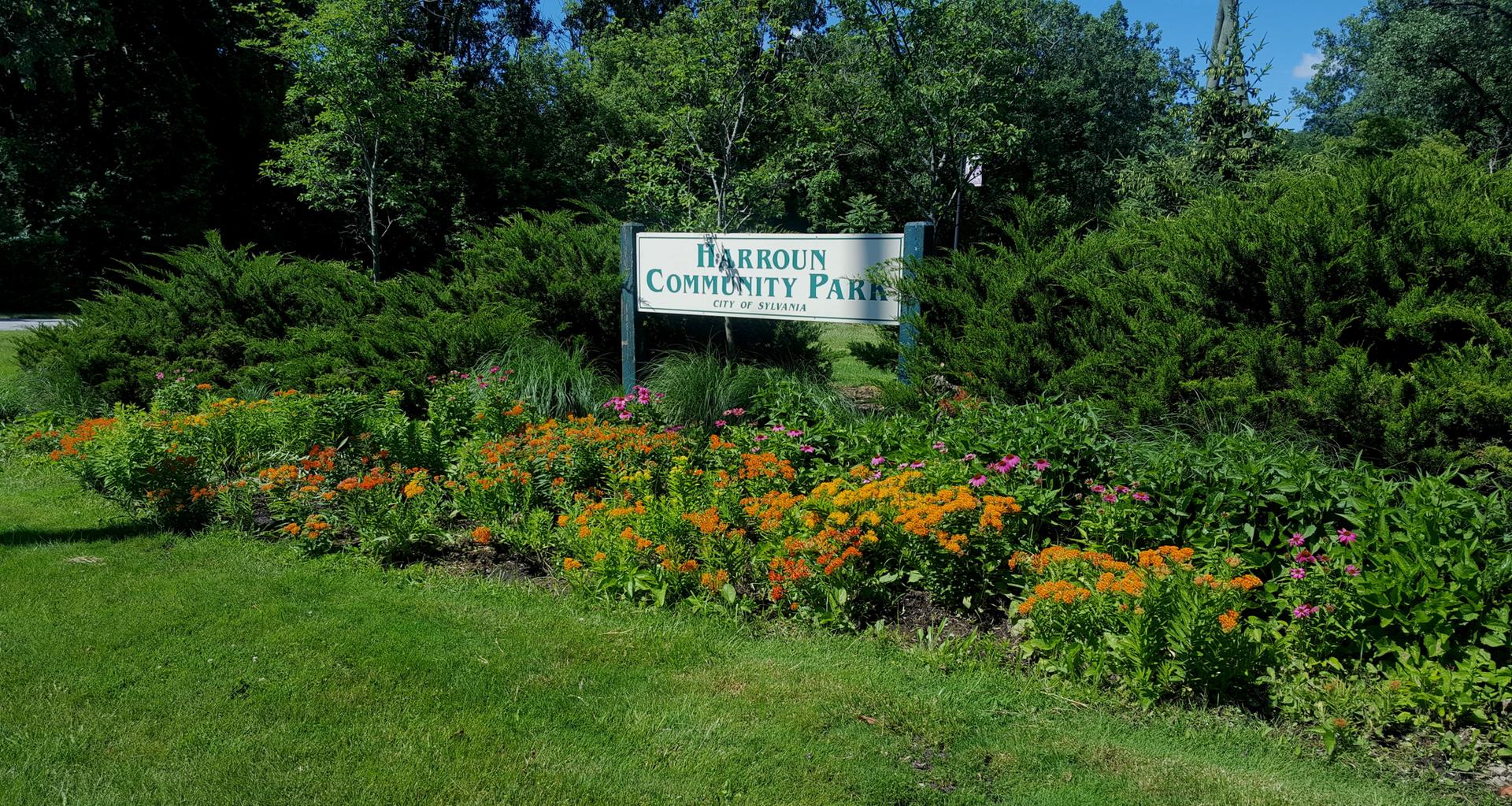 A park sign surrounded by vibrant flowers, green bushes, and trees on a sunny day.