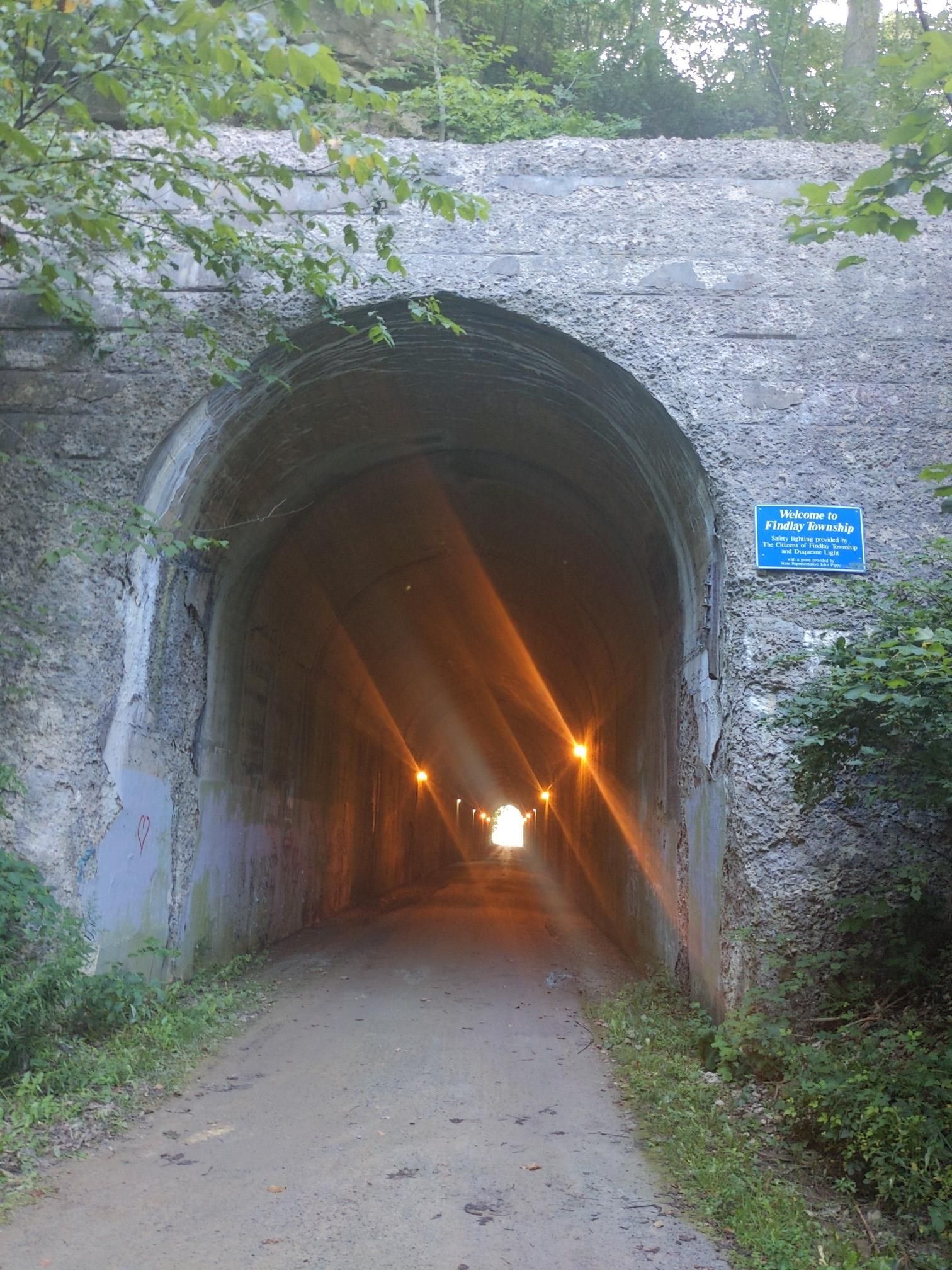 Arched stone tunnel entrance with a dirt path and bright interior lights. - Montour Trail