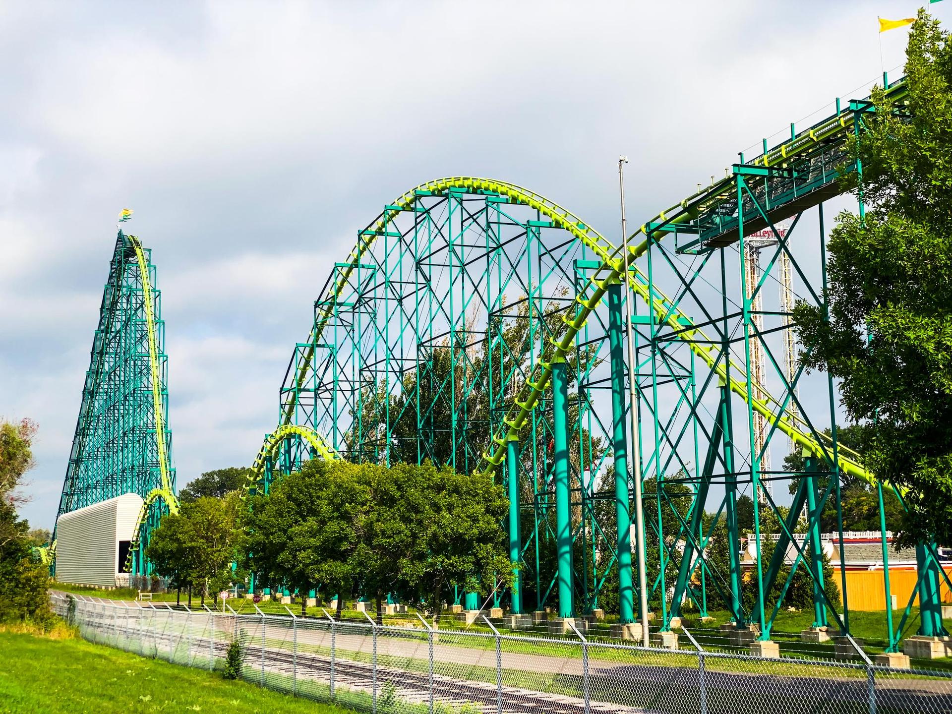 A towering green and blue roller coaster dominates the landscape under a cloudy sky.