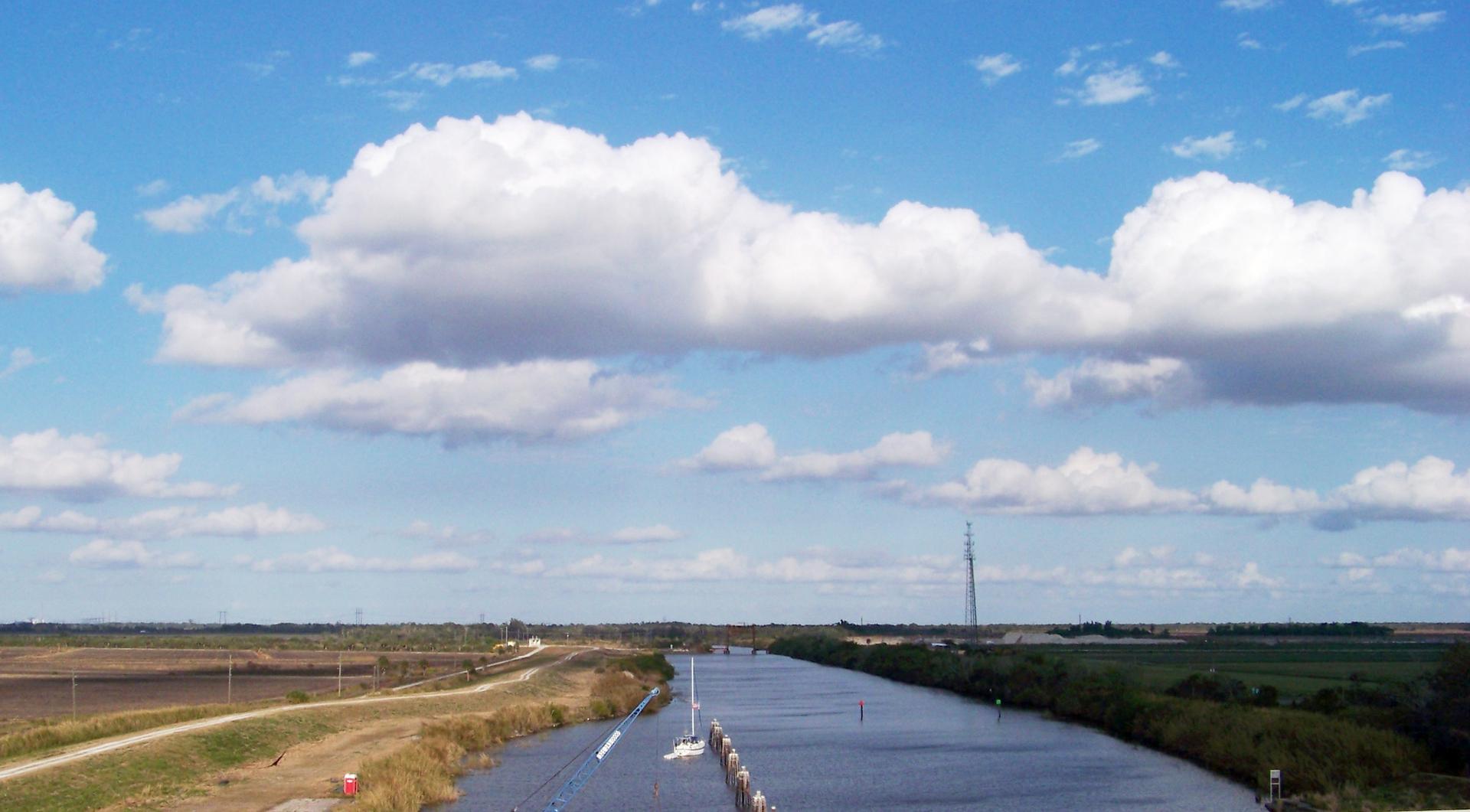 Wide canal flowing through flat fields under a bright, cloudy sky.