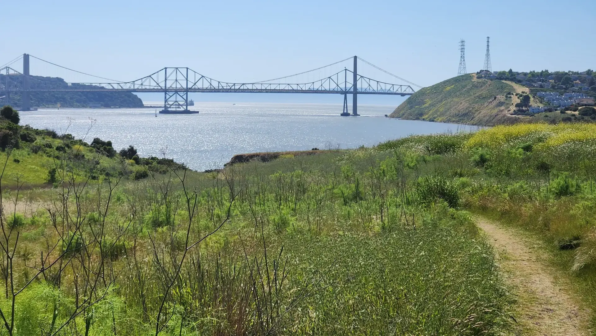 Dirt path through green grass leading to a large bridge over sparkling water.