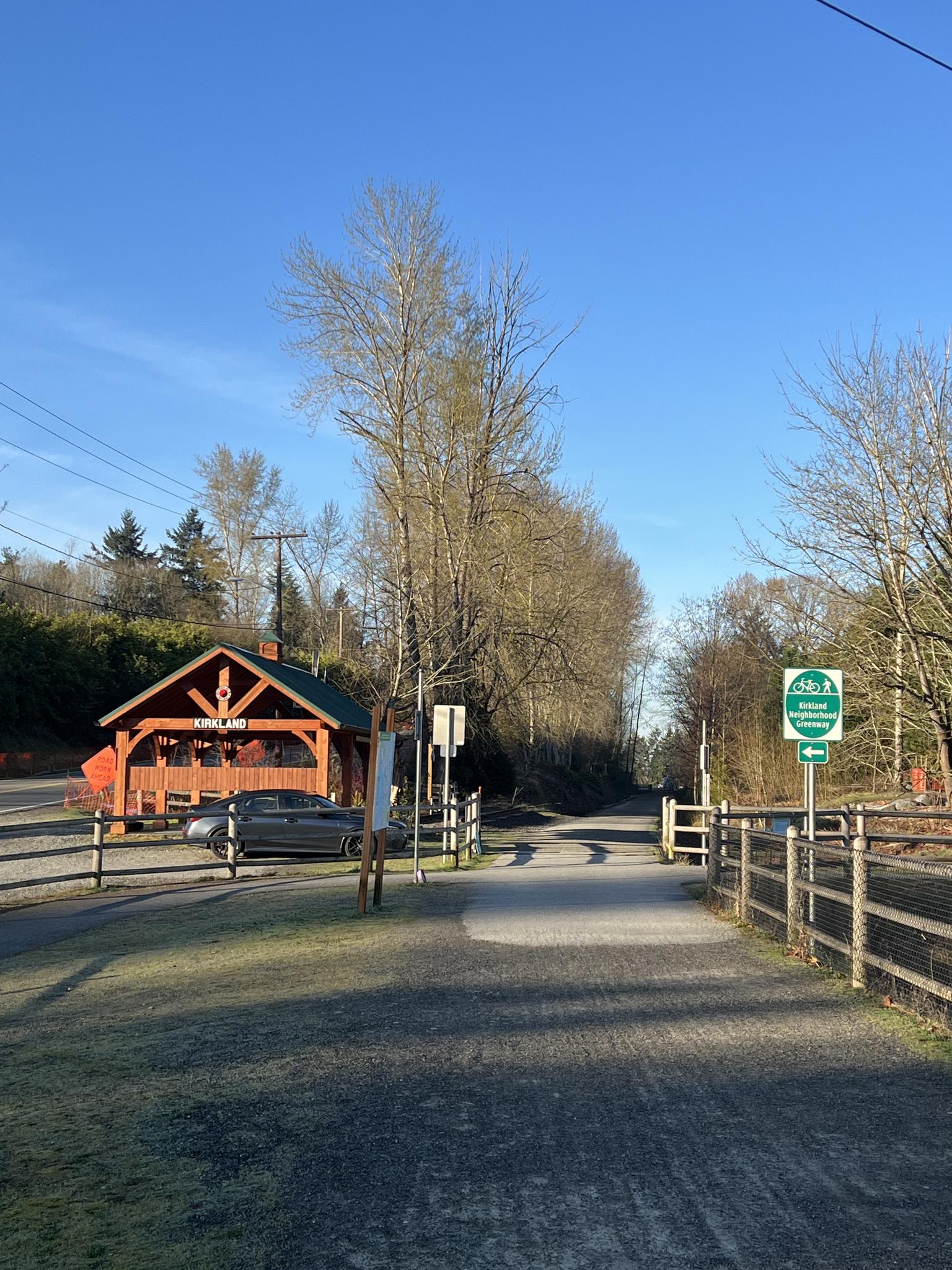 Sunny trail entrance with a rustic building, car, and bare trees.
