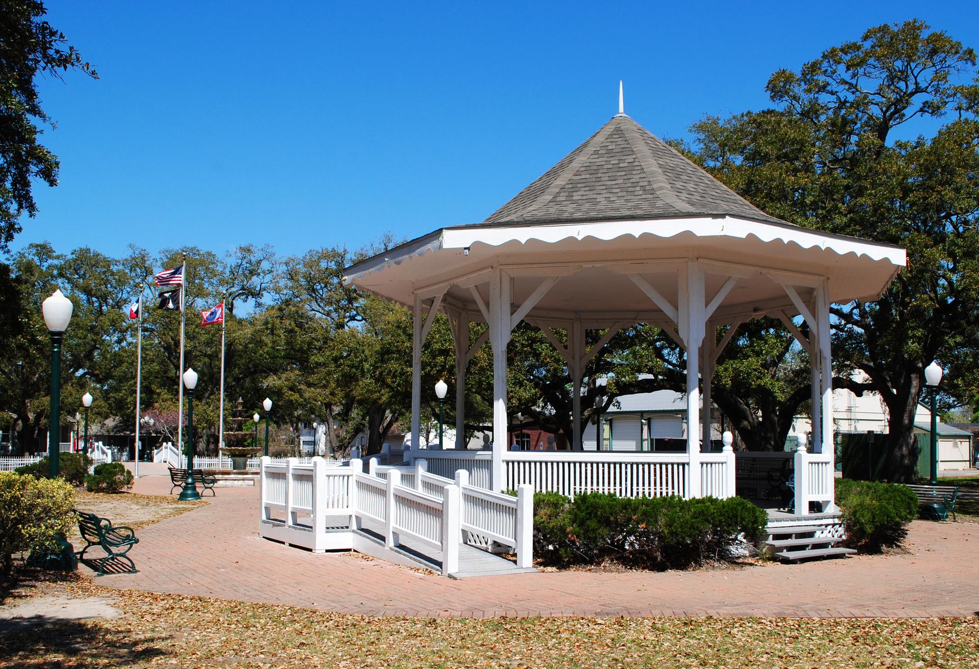 White gazebo with a dark roof in a sunny park with trees.