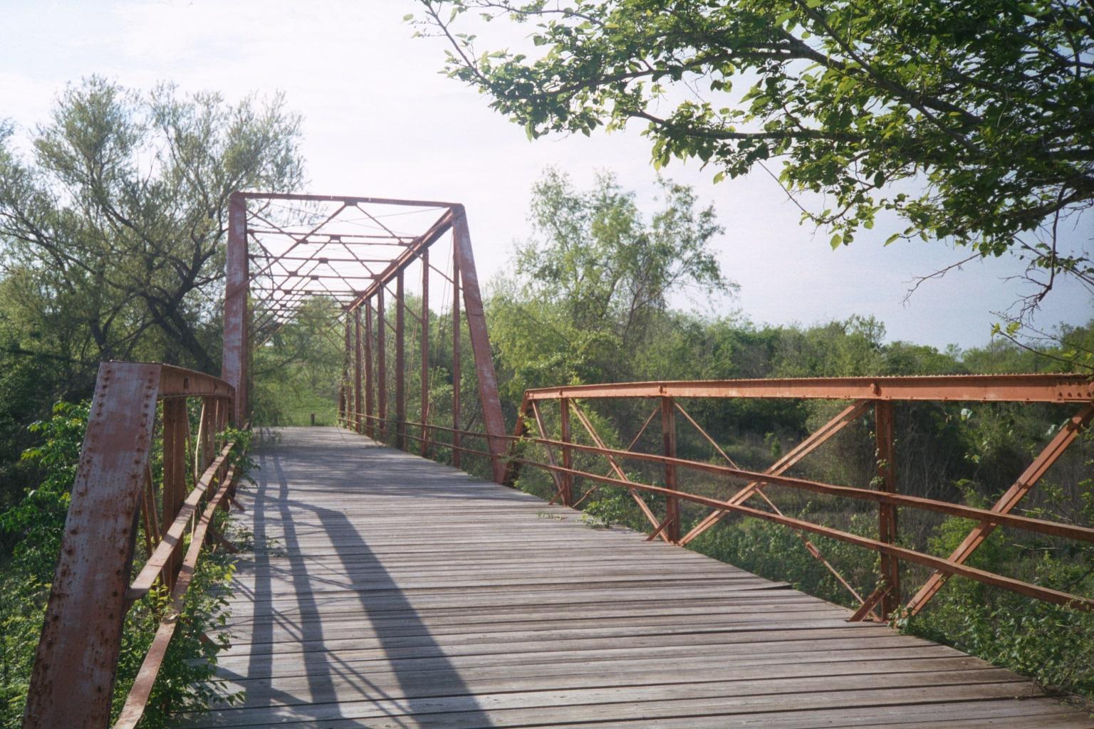 Rustic metal truss bridge with wooden deck, surrounded by green trees.