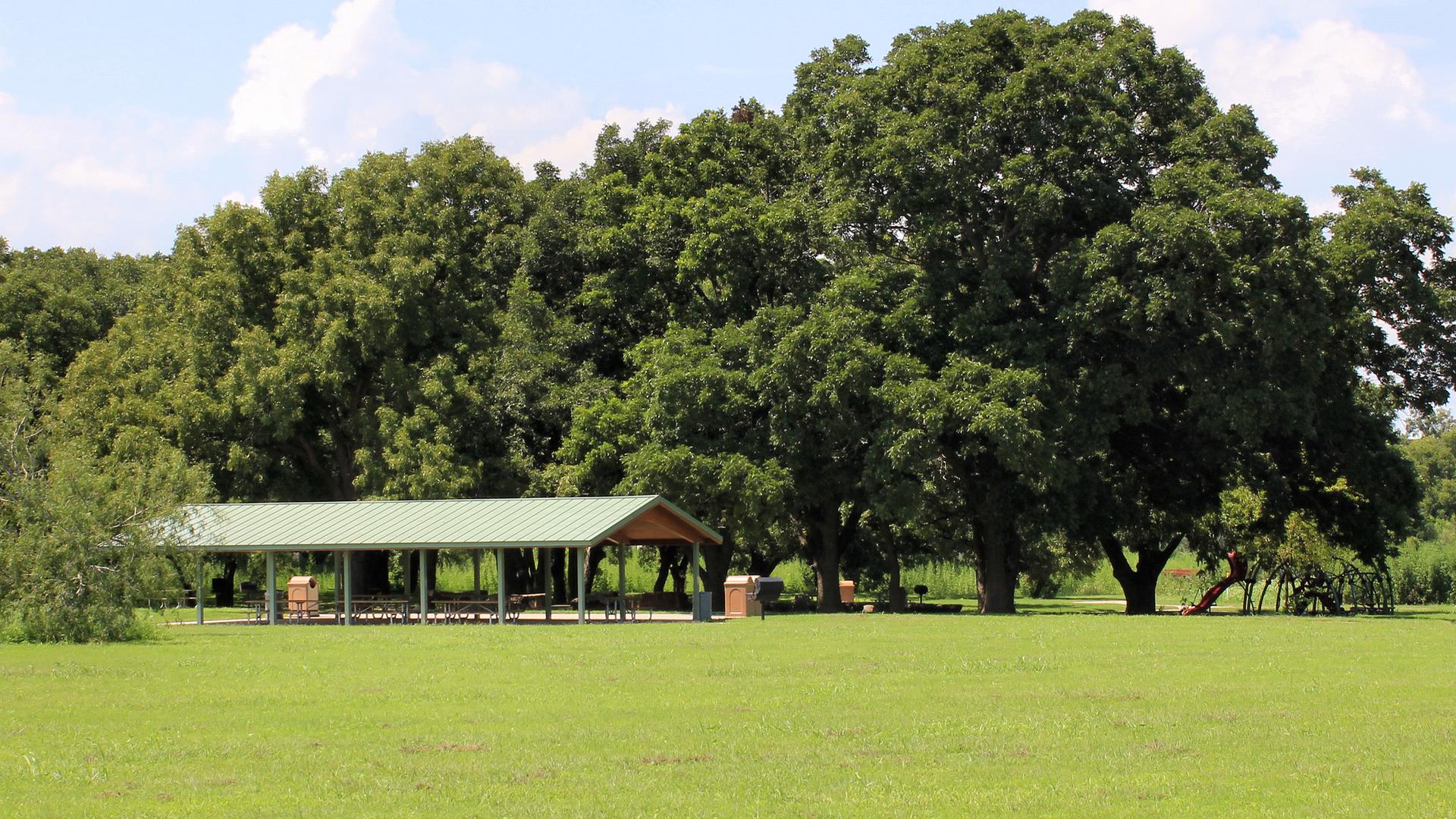 A green-roofed picnic pavilion sits in a sunny grassy field near trees.