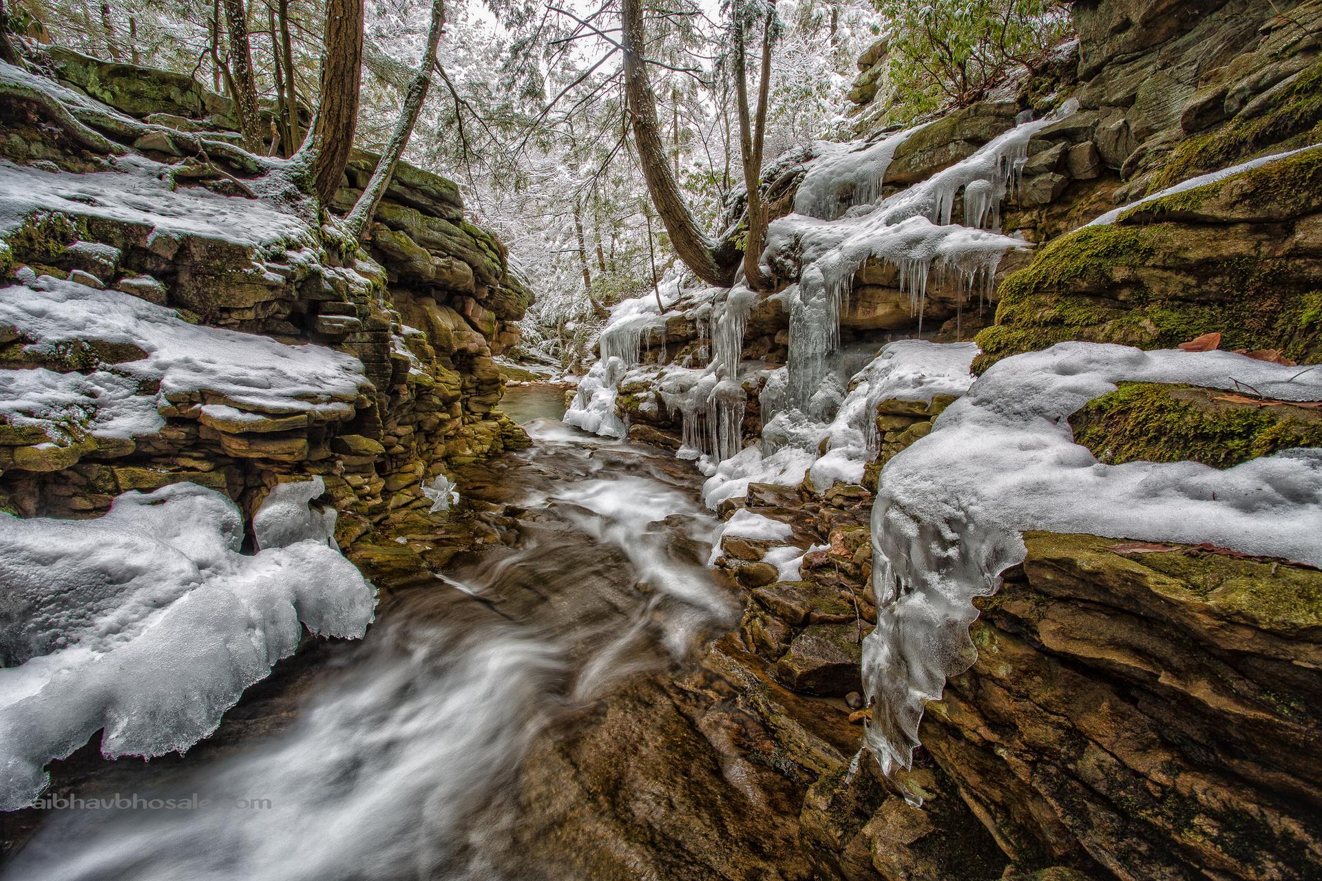A winter stream flows through an icy, snow-covered gorge with frosted trees.