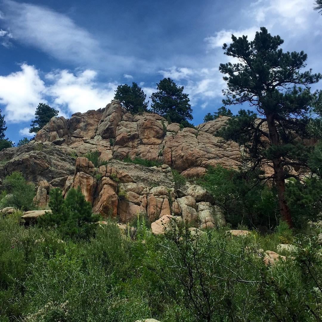 Towering rock formations on a hillside with green trees under a cloudy blue sky.