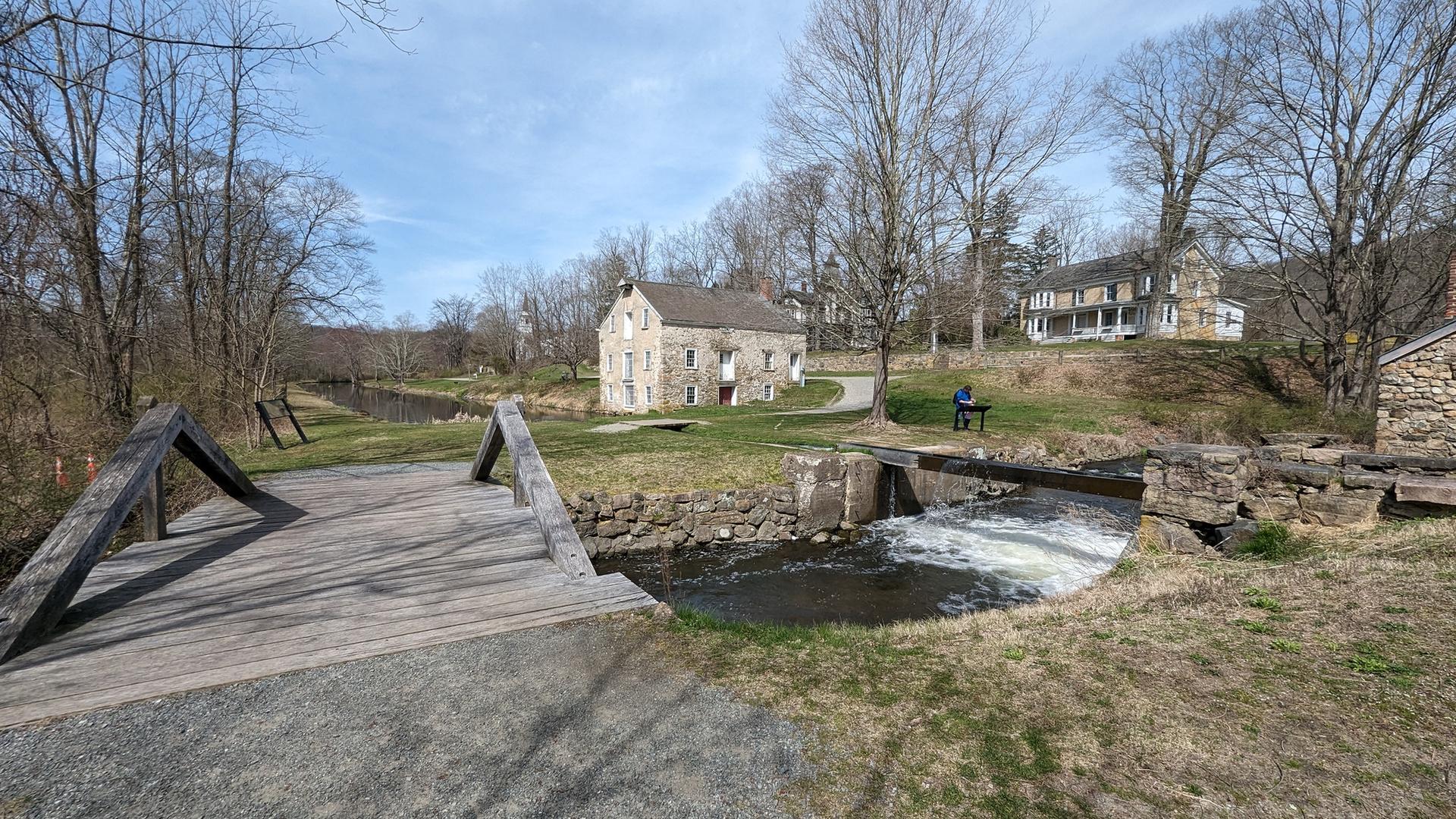 Historic stone building by a river with a wooden bridge and person sitting.