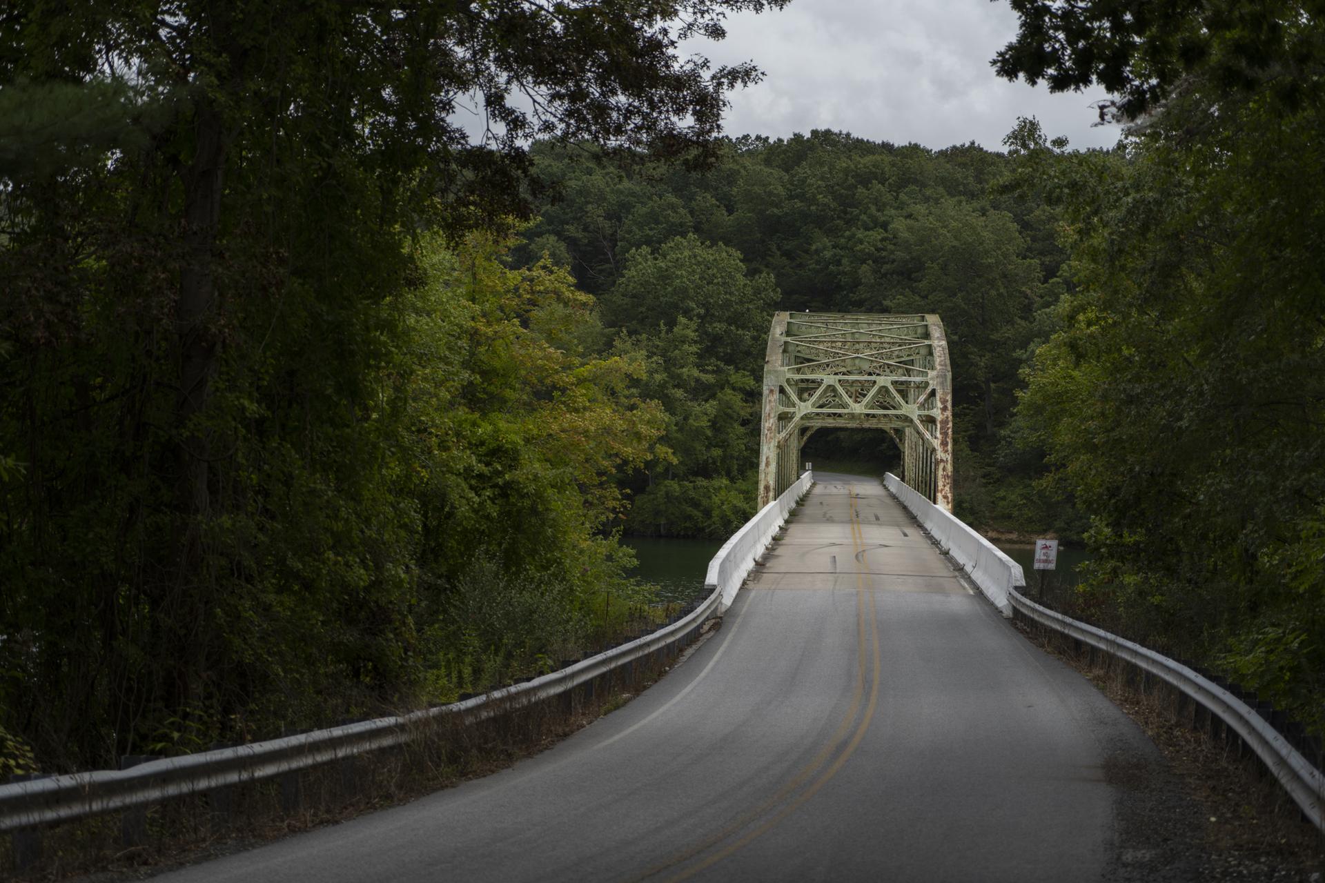 Road leading to an old green truss bridge through dense, dark forests.