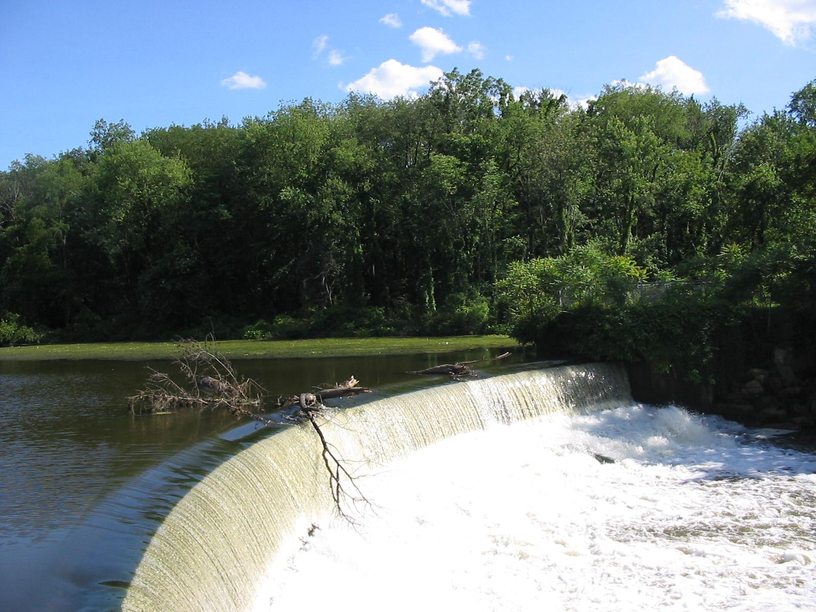 A wide waterfall flows into a river, surrounded by lush green forest under blue sky.