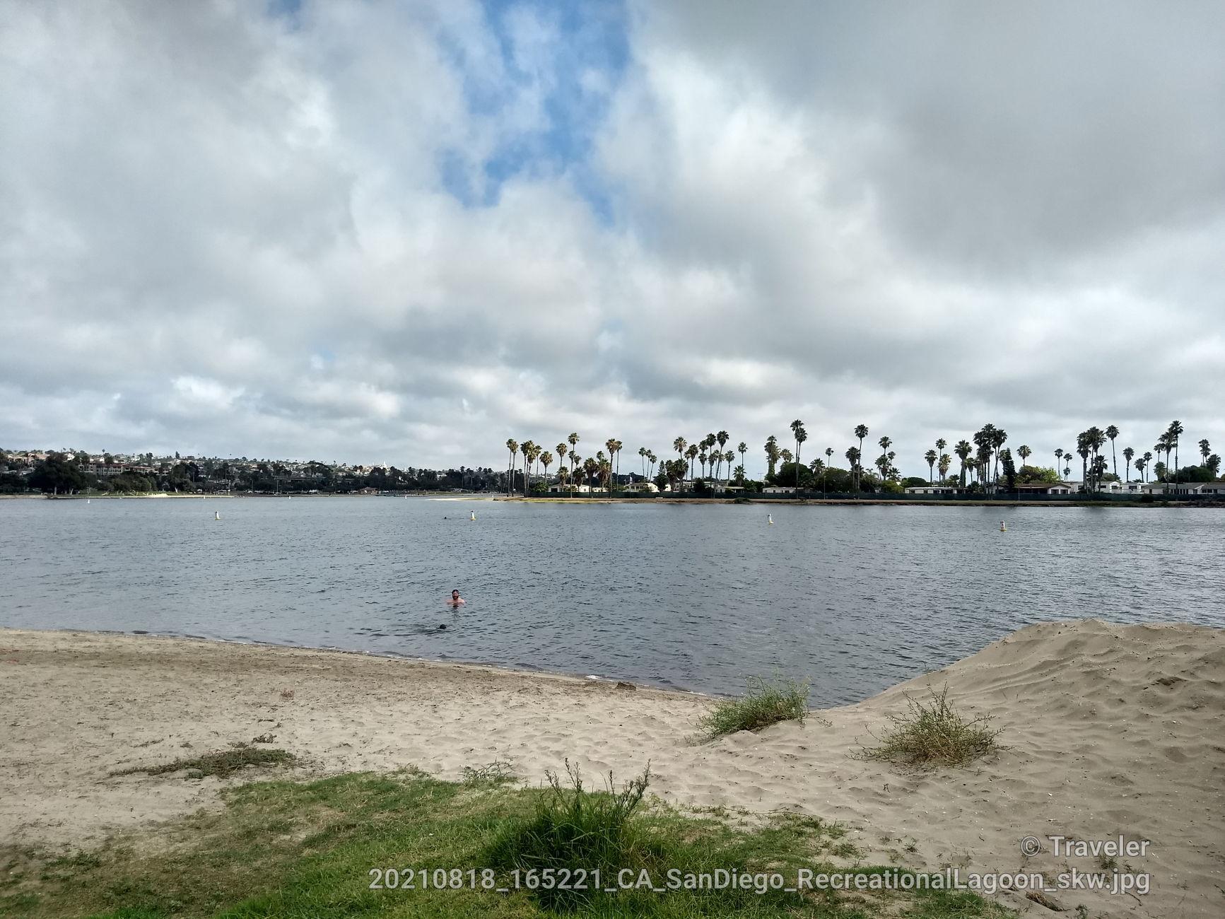 Person wades in calm water near a sandy beach under cloudy sky.