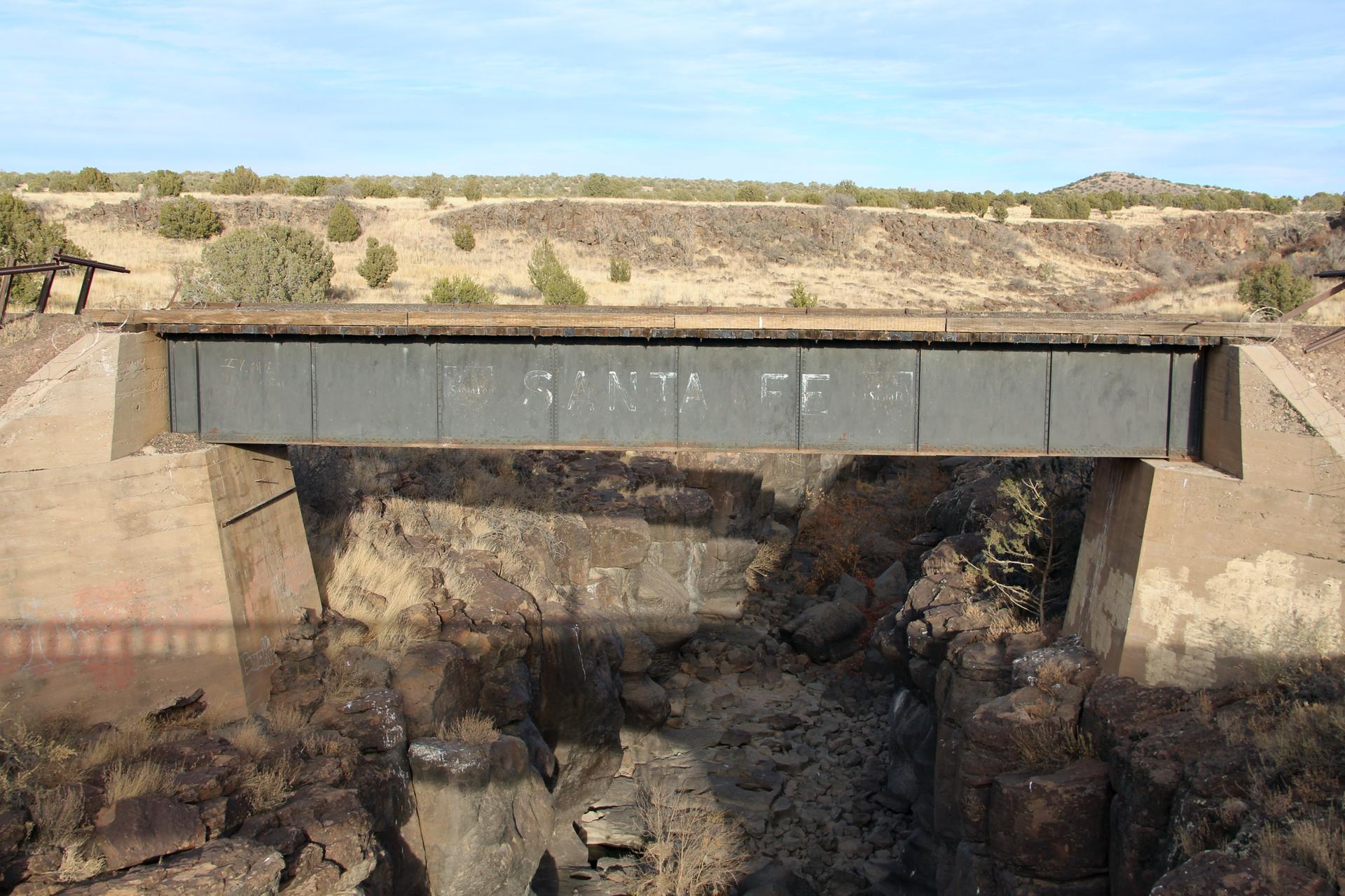 A metal and concrete bridge spans a dry, rocky canyon under bright daylight.