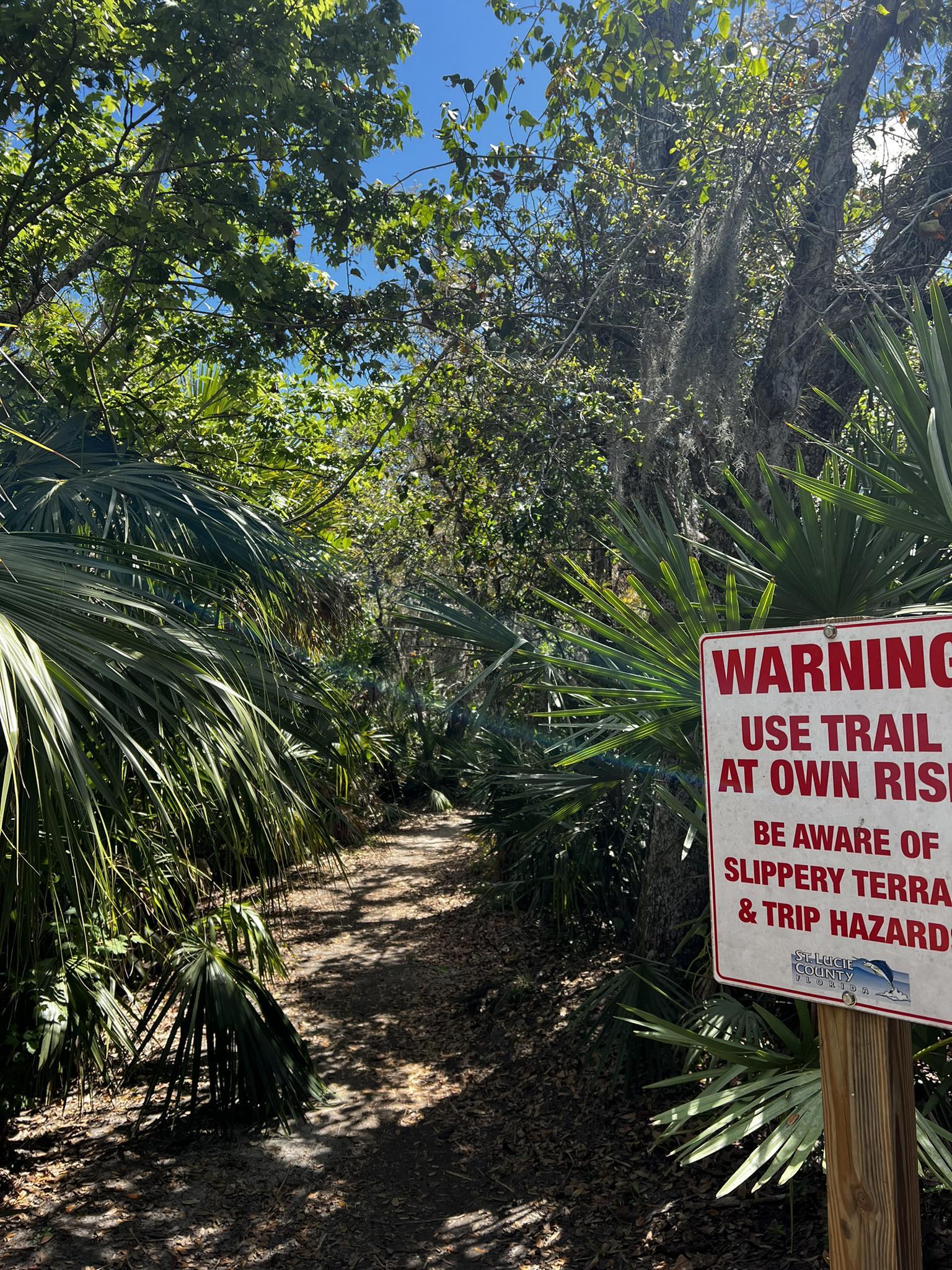Warning sign beside a shaded, narrow dirt trail through dense green foliage.