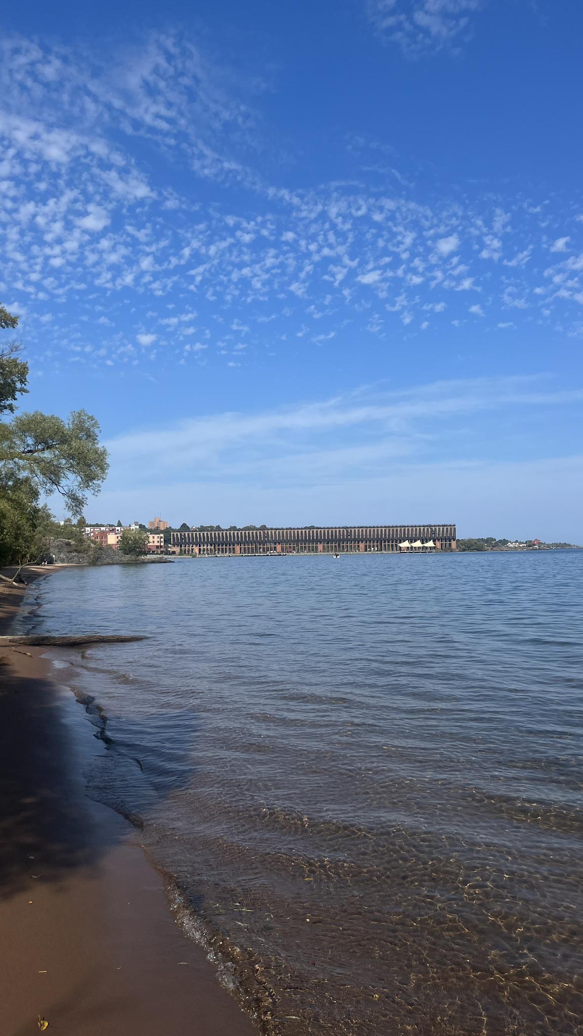 Calm water laps a sandy beach with a distant industrial building under blue sky.