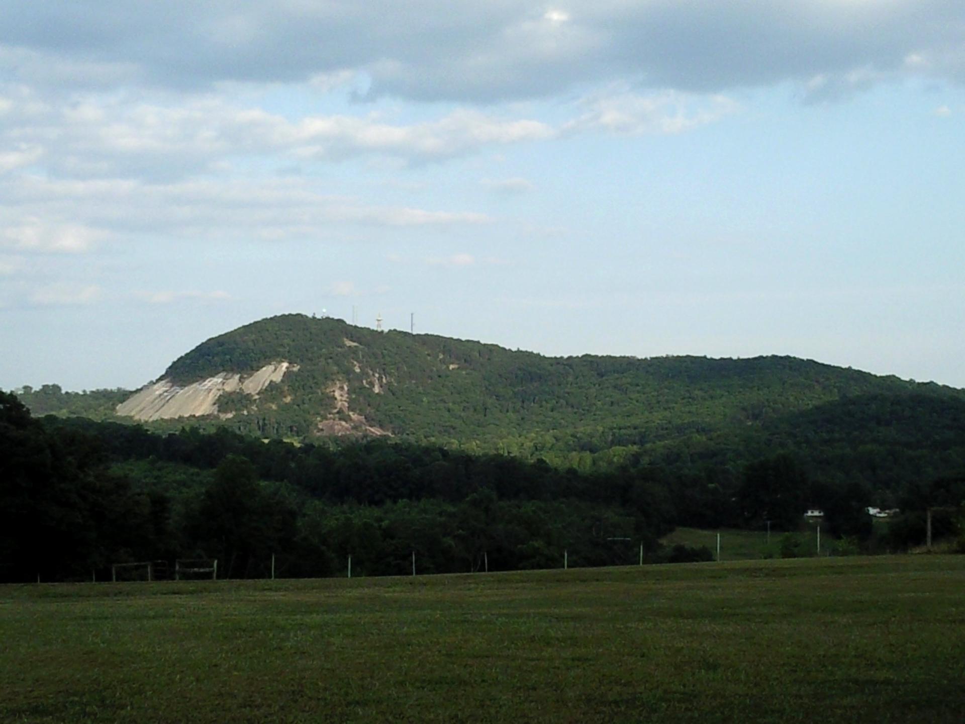A large, green mountain with a rocky face overlooks a grassy field under a cloudy sky.