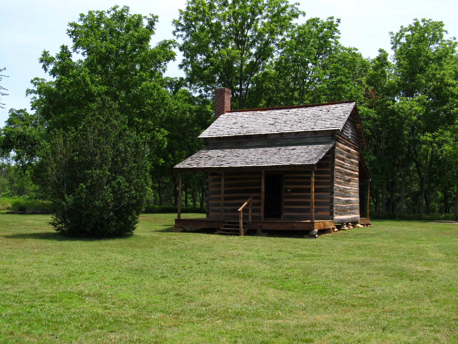 Rustic log cabin with a porch, surrounded by green grass and trees.