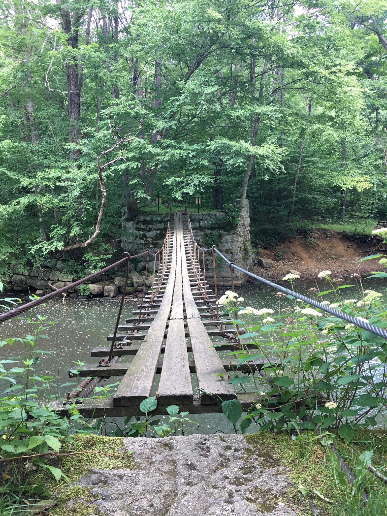 A rustic wooden suspension bridge spans a river in a lush green forest.
