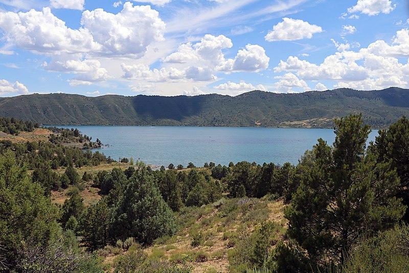 Bright day over a blue lake surrounded by green trees and distant mountains.