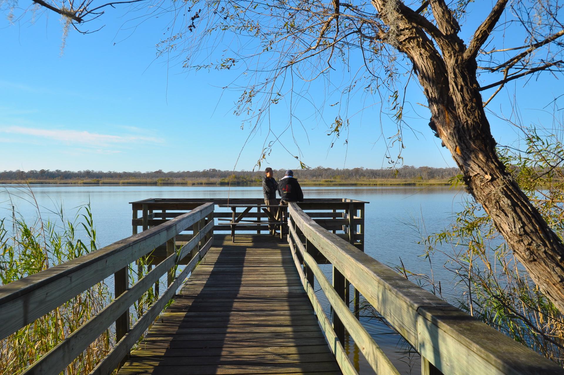 Two people sit on a wooden dock overlooking a wide lake.