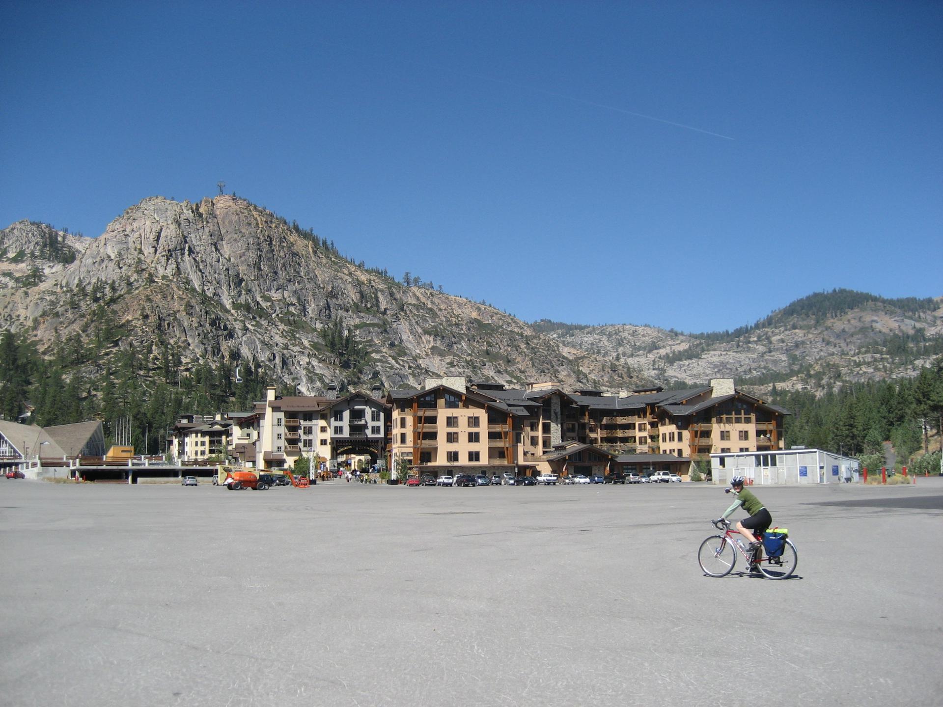 A cyclist rides across a large paved area with buildings and rocky mountains.