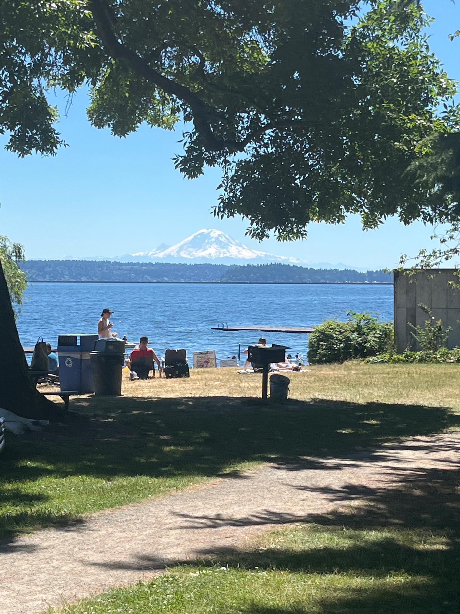 Two people by a sunny lake with a distant snow-capped mountain under blue sky.