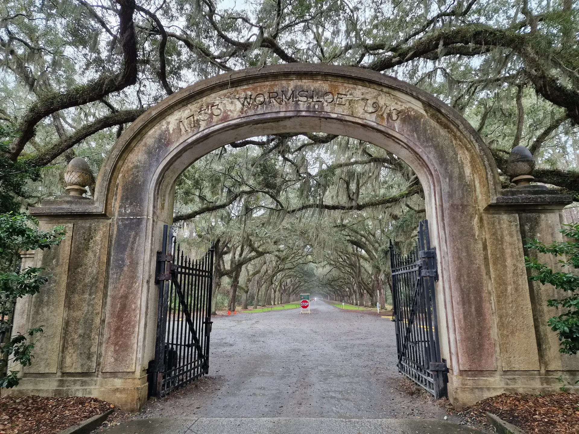 Ornate stone archway with open gates leading to a long, tree-lined road.