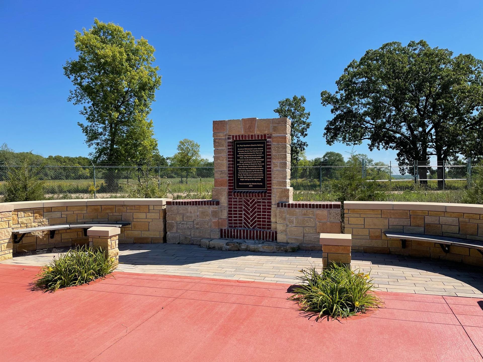 A stone monument with benches on a red paved area under a clear blue sky.