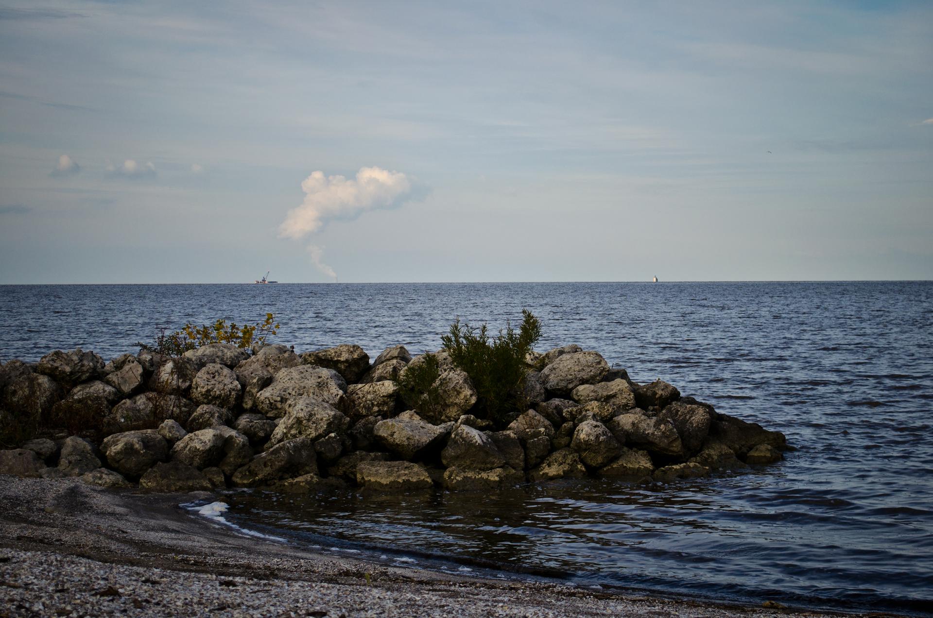 Rocky outcrop in calm water under a cloudy sky, with distant plume.