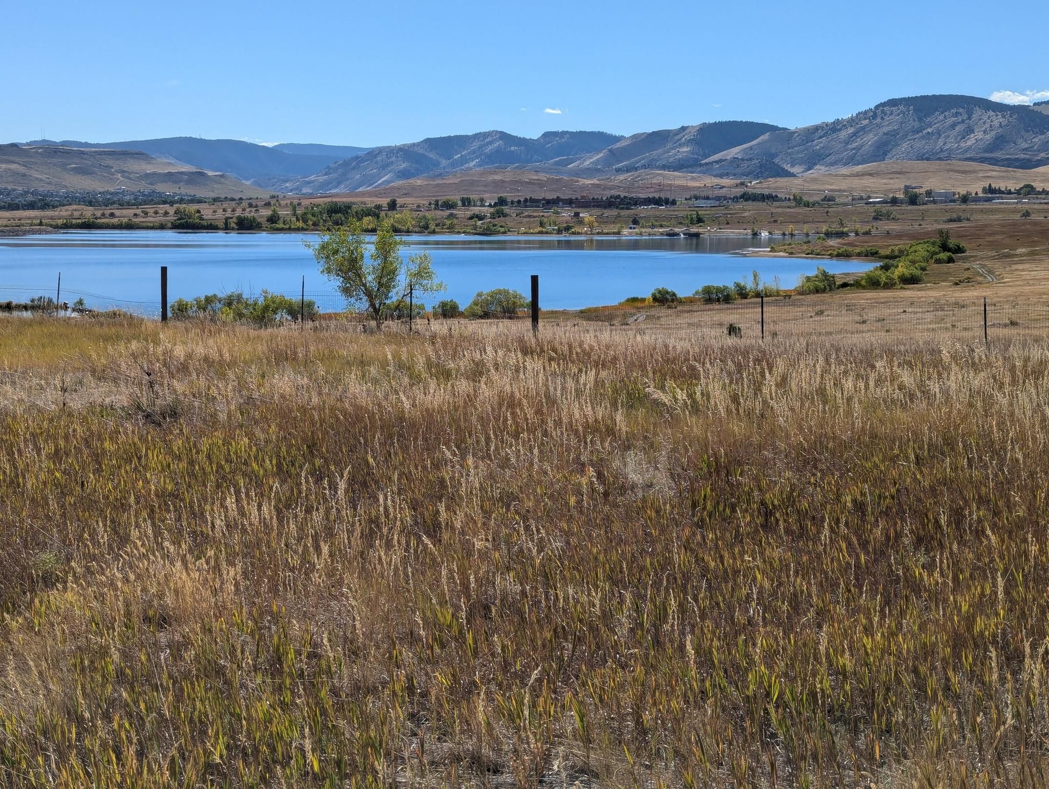 A calm lake surrounded by dry grass and distant mountains under a clear sky. - View of the Arvada Blunn Reservoir – Tucker Lake View loop from Arvada West Dog Park