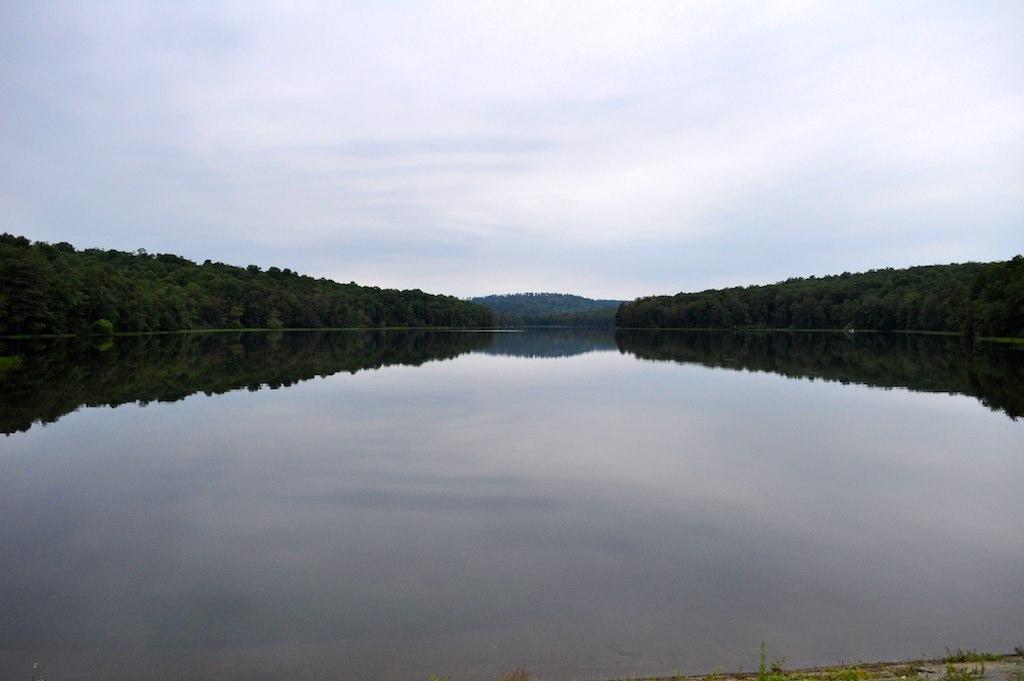 A calm lake reflects forested hills under an overcast sky.