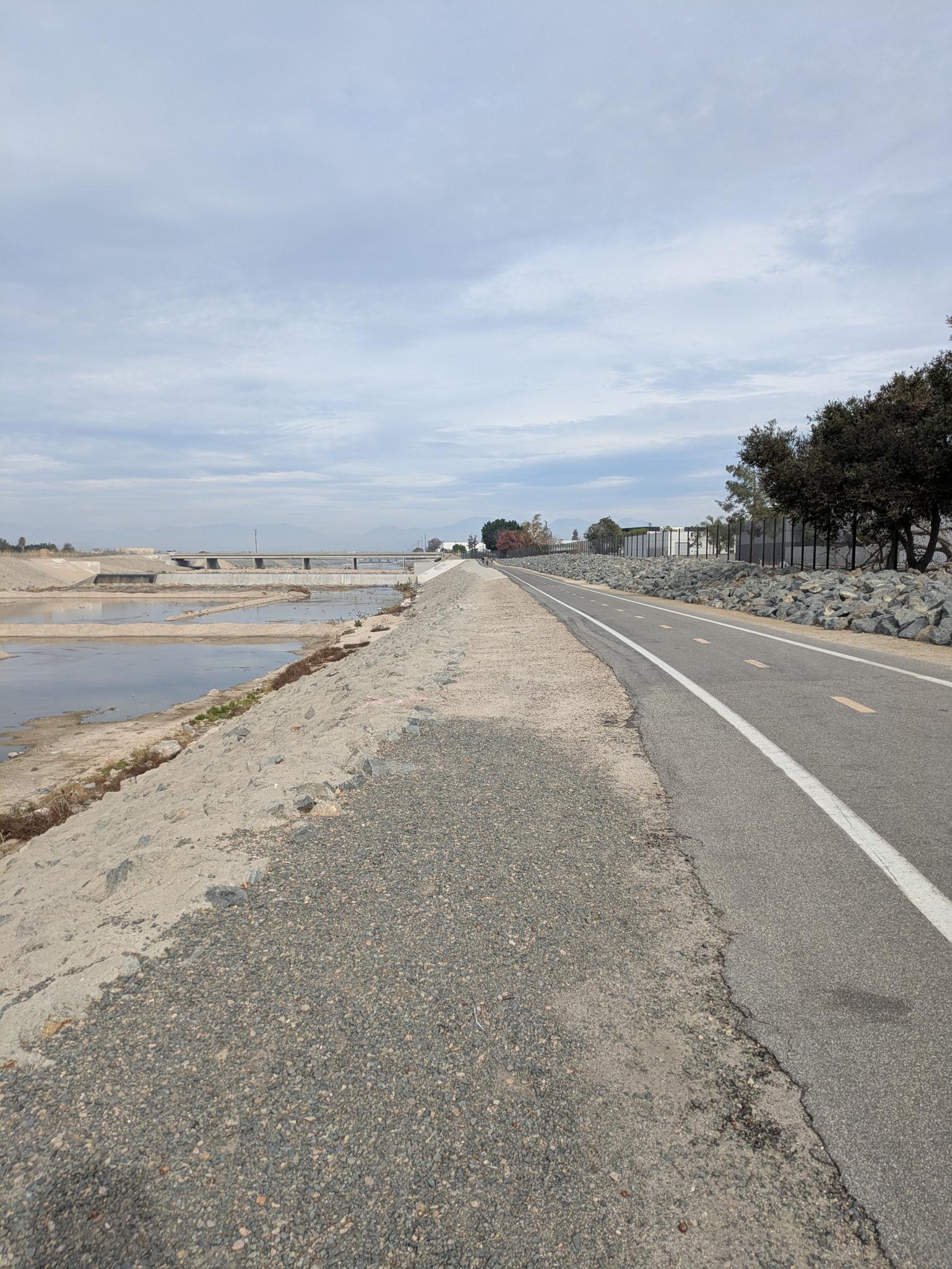 A paved path and gravel trail next to calm water under an overcast sky.