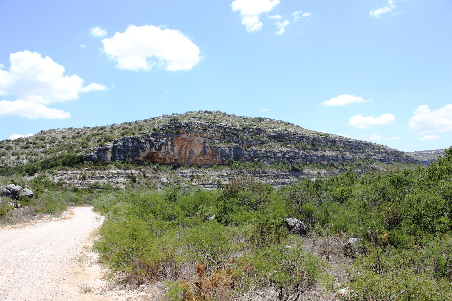 Dirt road leading towards a rocky hill with visible cliffs and green trees.