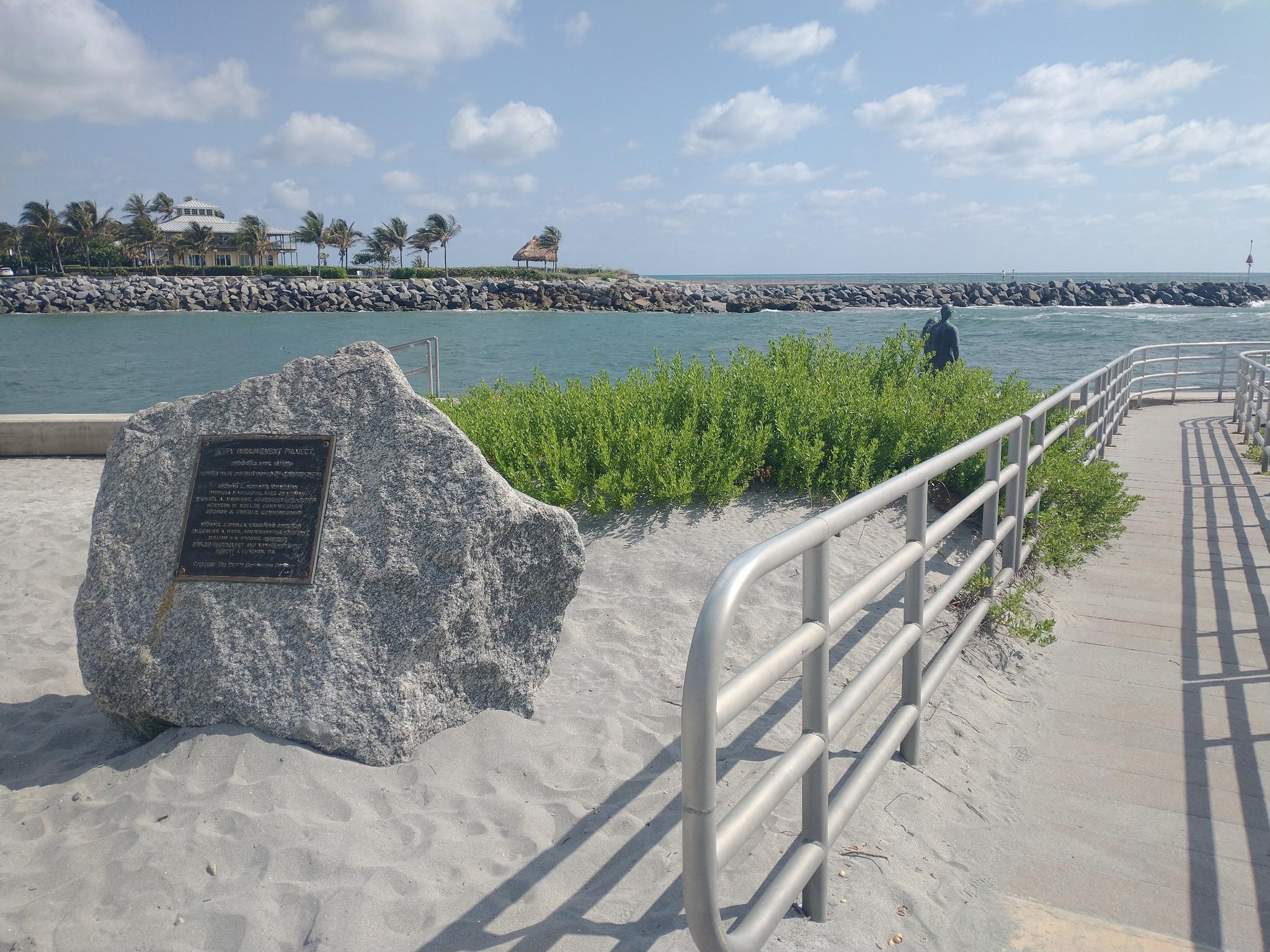 A large rock with a plaque sits on a sandy path near a water jetty.