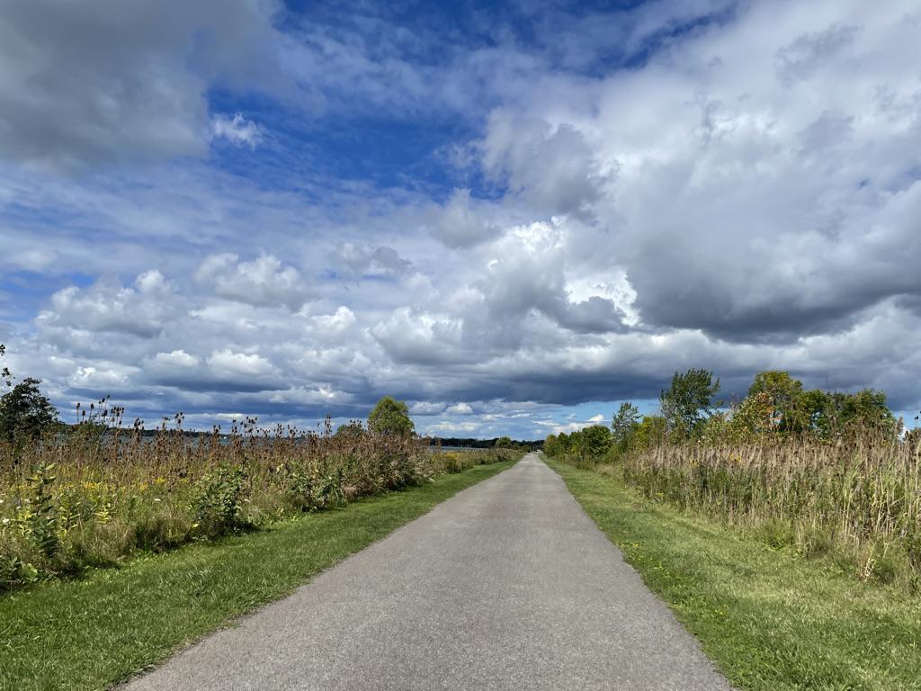 A paved path stretches through green fields under a bright, partly cloudy sky.