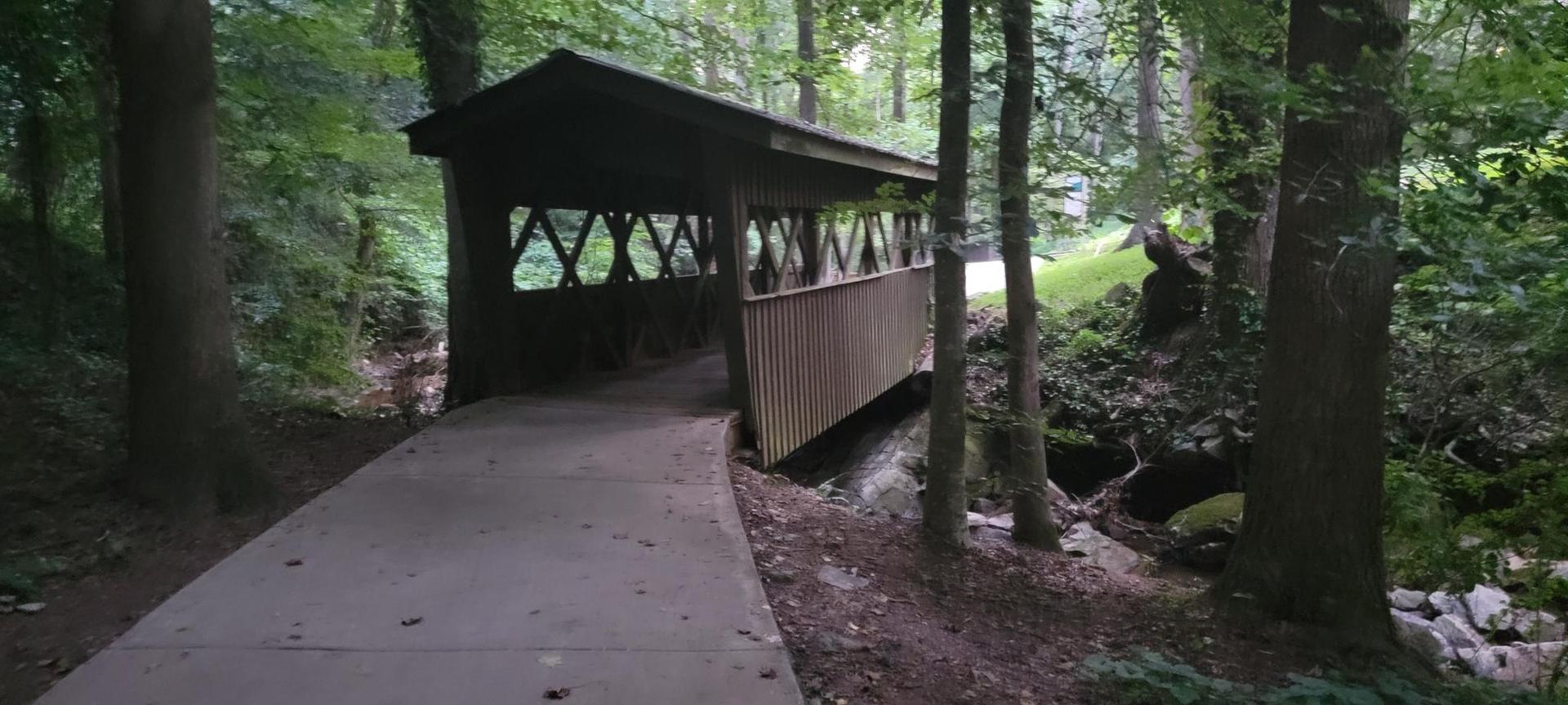 Covered wooden bridge over a stream in a shaded, green forest.