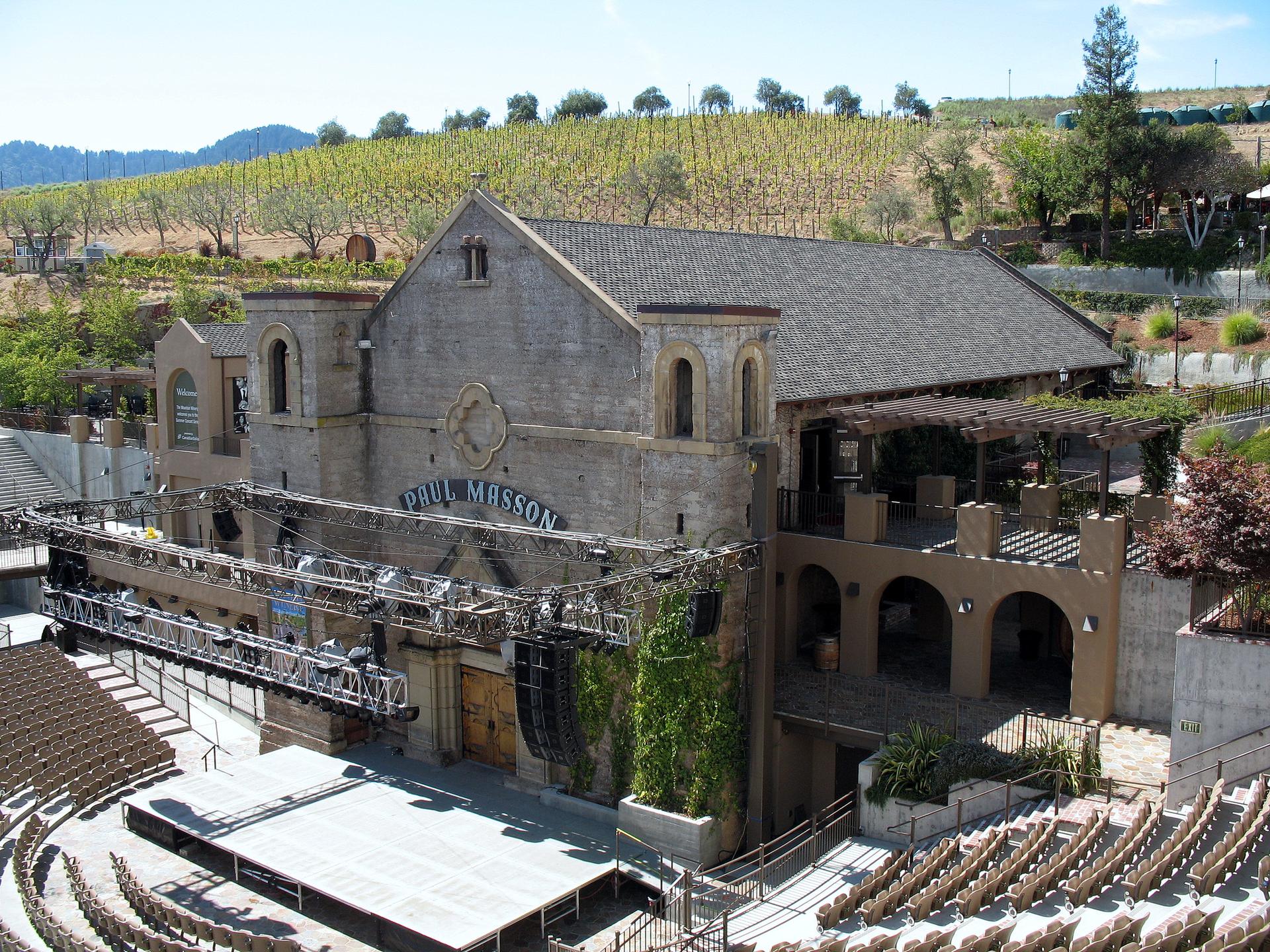 A grand stone building with an outdoor amphitheater and vineyards on hills.